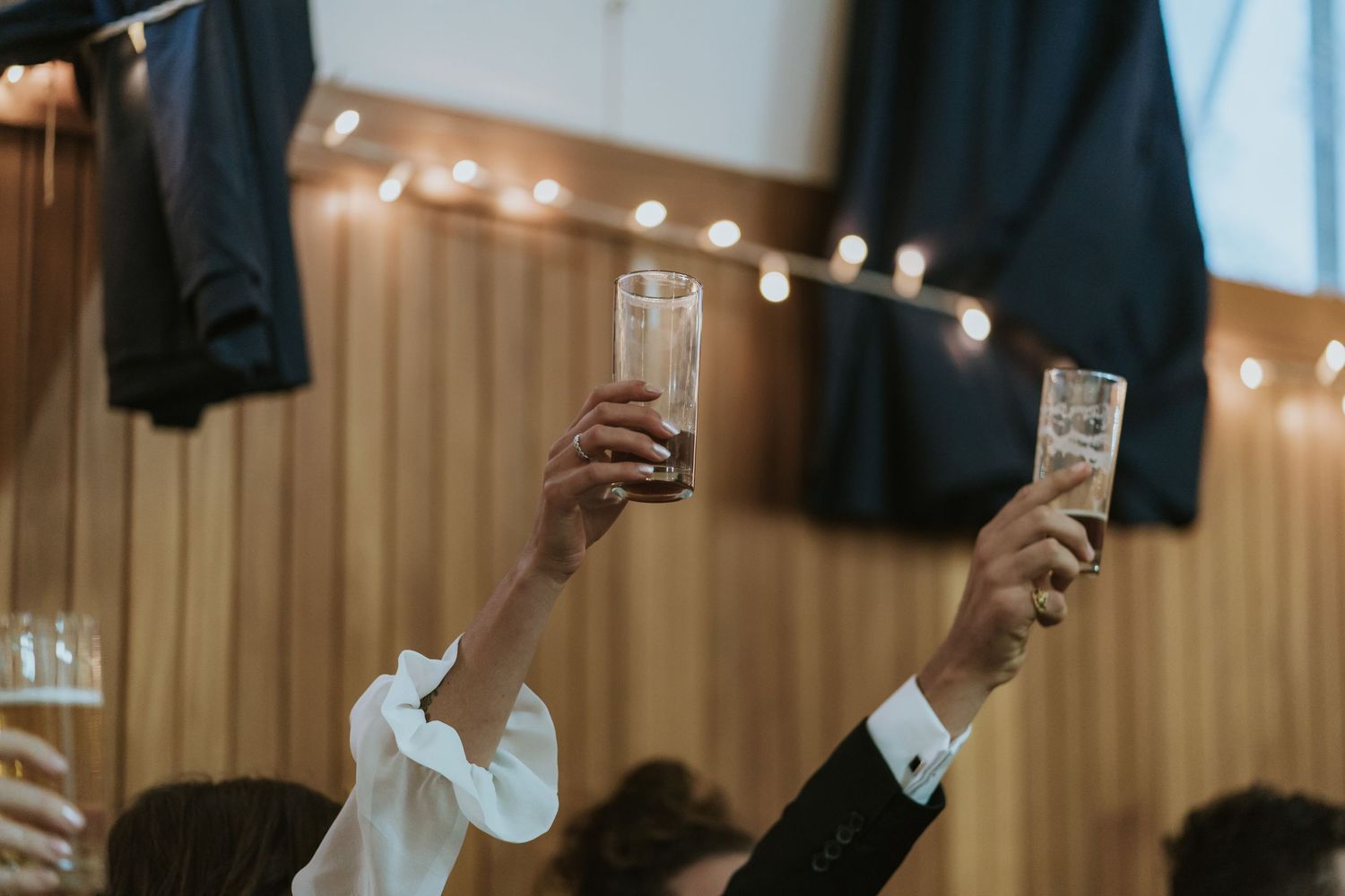 People raise glasses in celebration with string lights glowing in the warm wooden interior background.