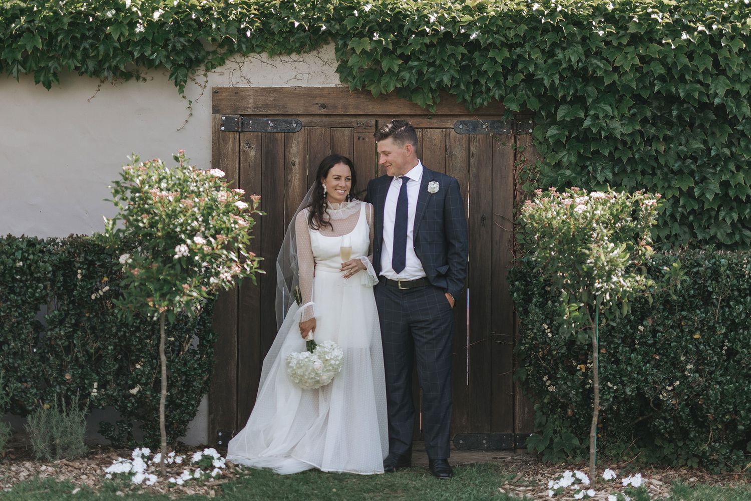 Bride and groom share a romantic moment in garden setting with green ivy wall and rustic wooden door backdrop.