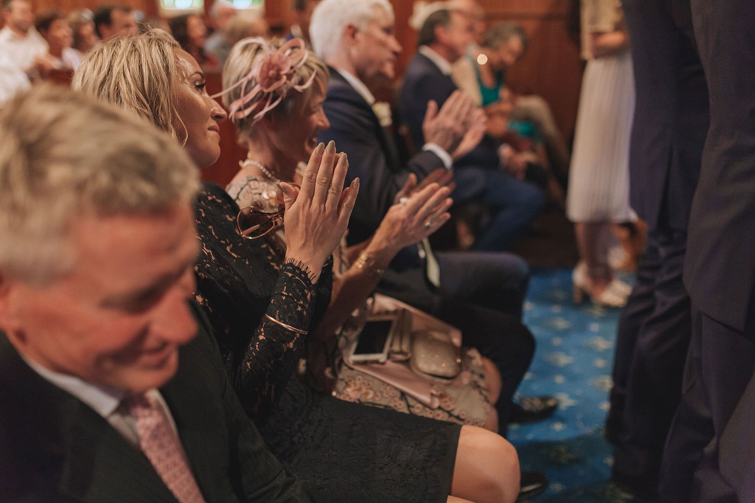A crowd of seated people applauding during an event in an auditorium with blue carpet and dim lighting.