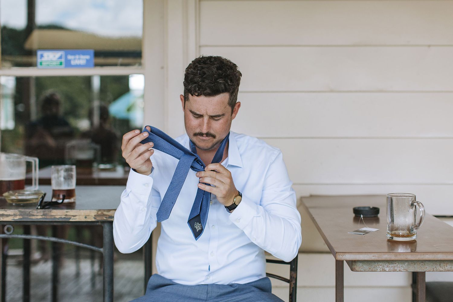 Someone in a crisp white dress shirt adjusts a blue necktie while seated at an outdoor cafe table.