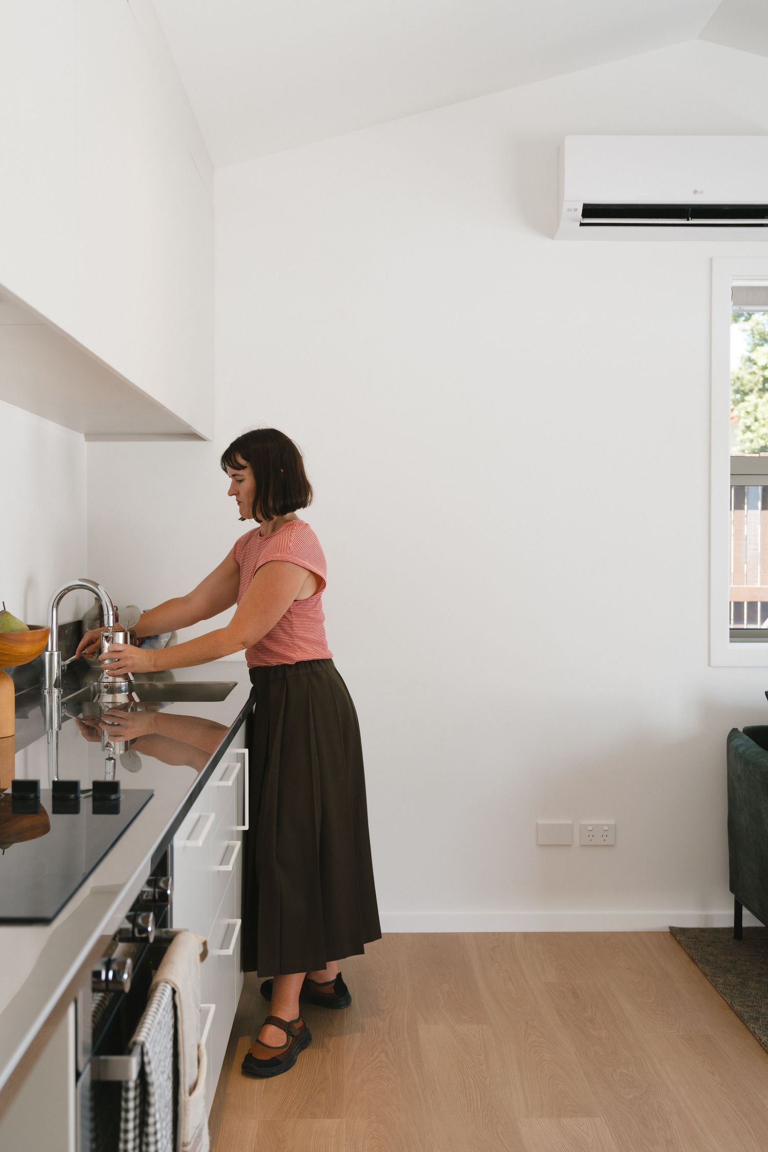 Woman in pink top and dark skirt filling a glass at a modern kitchen sink with white walls and timber floors.