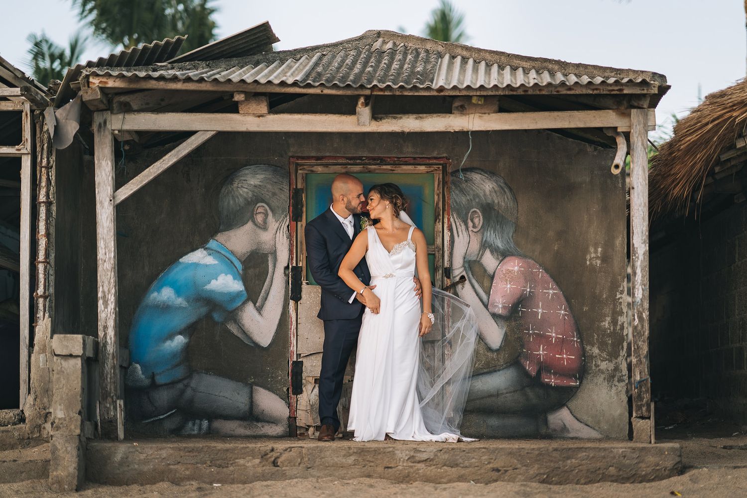 A couple in wedding attire poses in front of a street art mural showing photographers taking pictures on a wall.