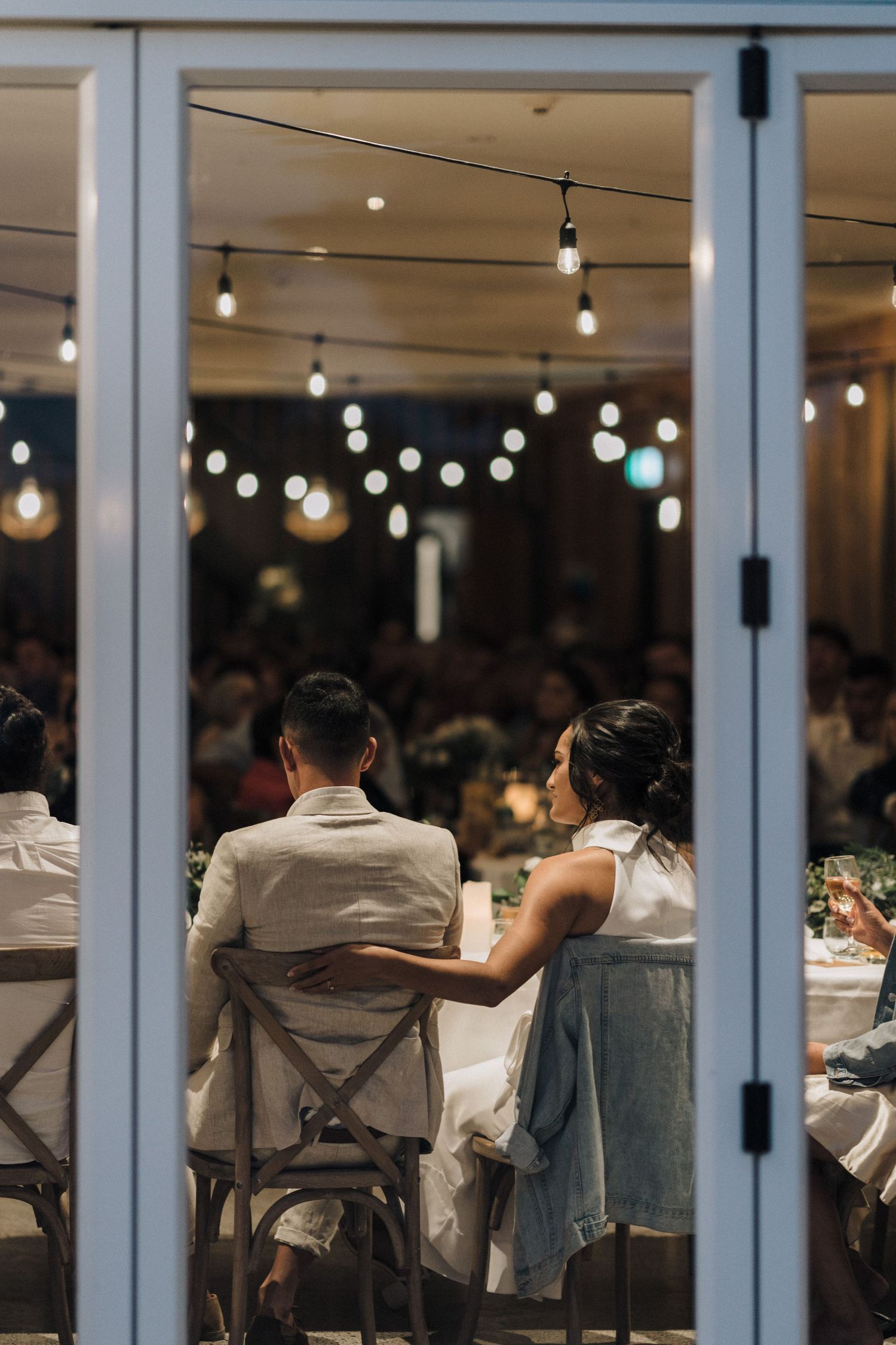 Wedding reception venue with string lights and guests seated at tables viewed through glass doors.