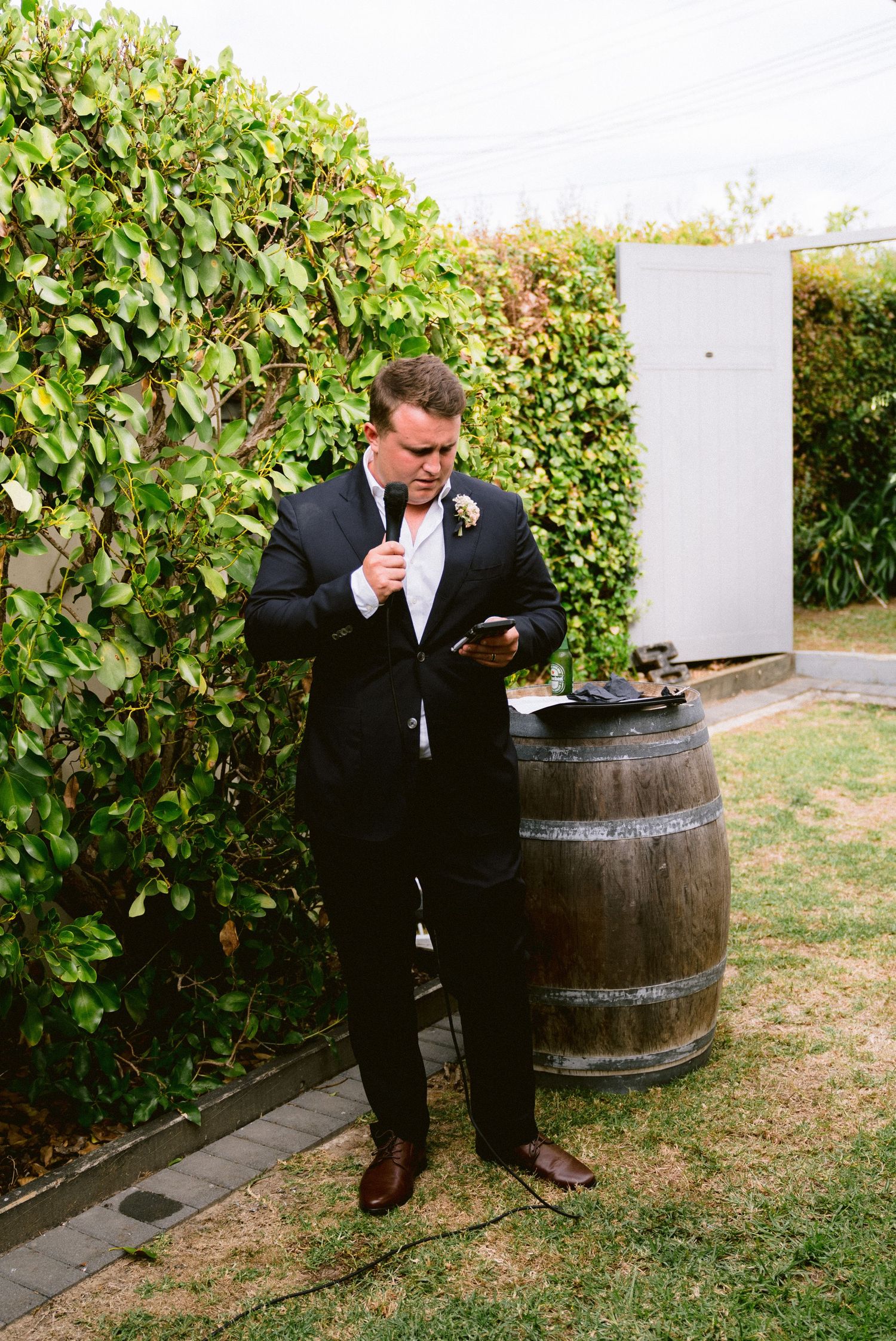 Groomsman in black suit checking phone while standing next to rustic wine barrel outdoors.