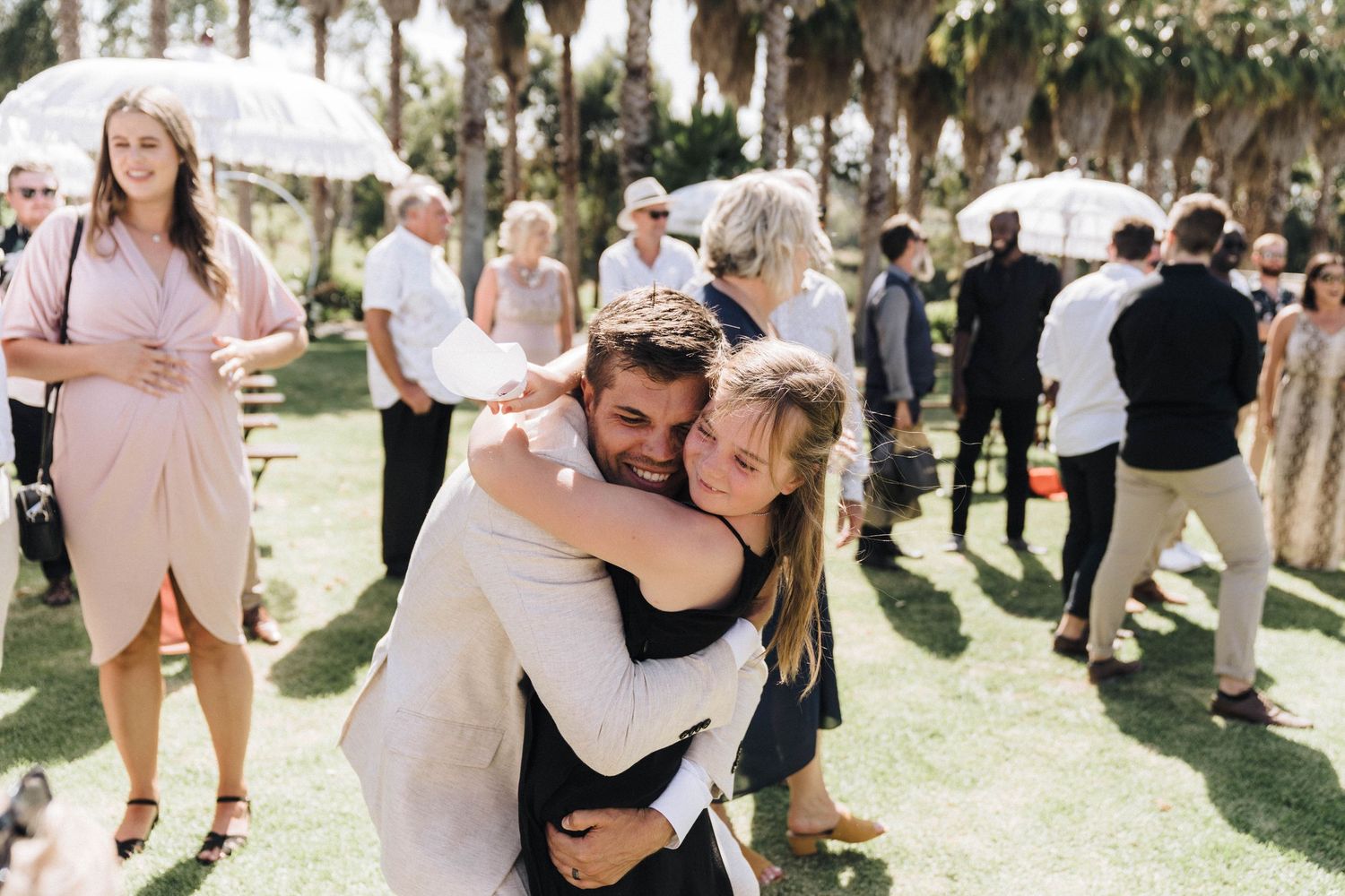 Happy couple embraces during outdoor wedding reception with guests mingling in background.