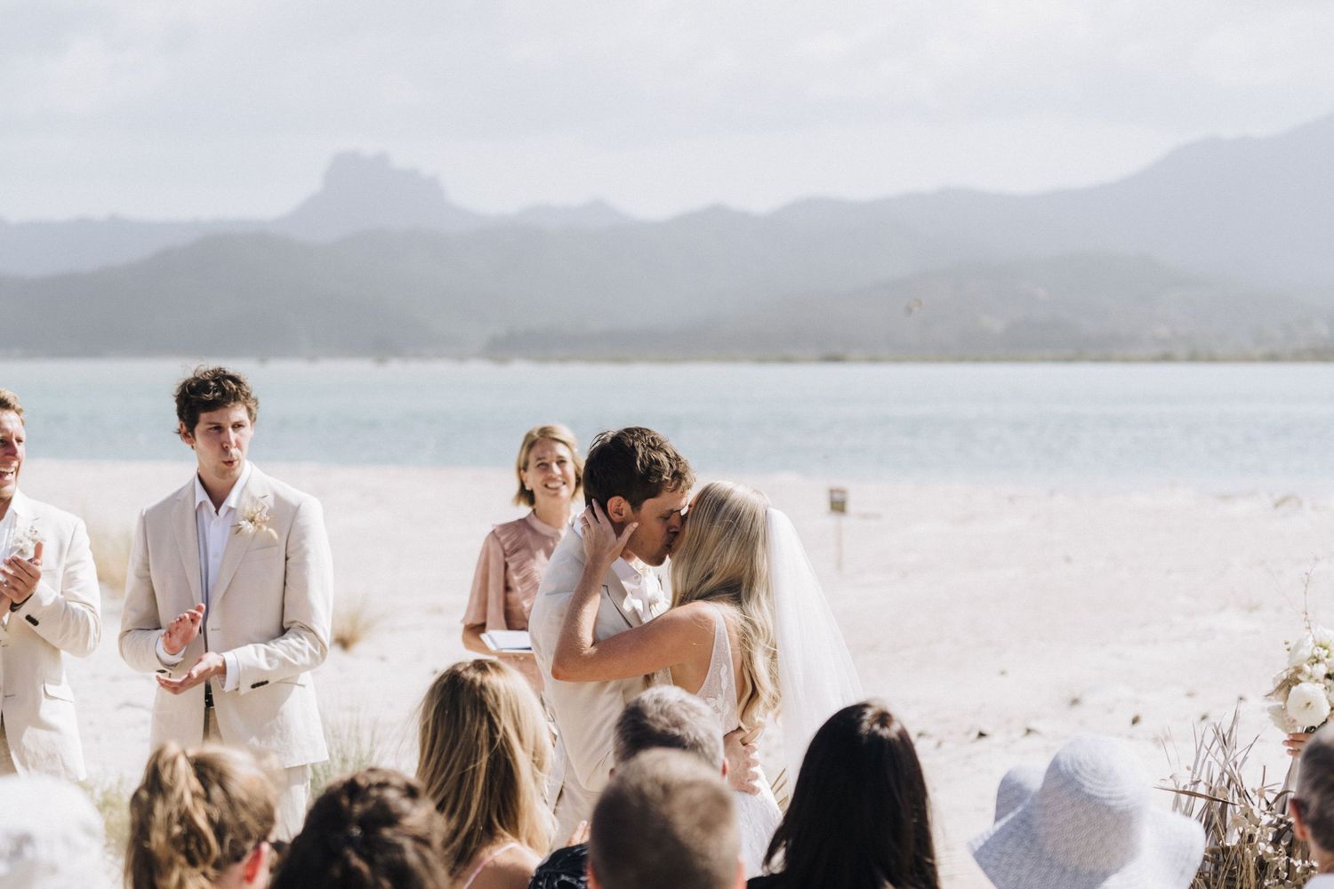 Seaside wedding panorama showing guests gathered on white sand with mountains in the distance.