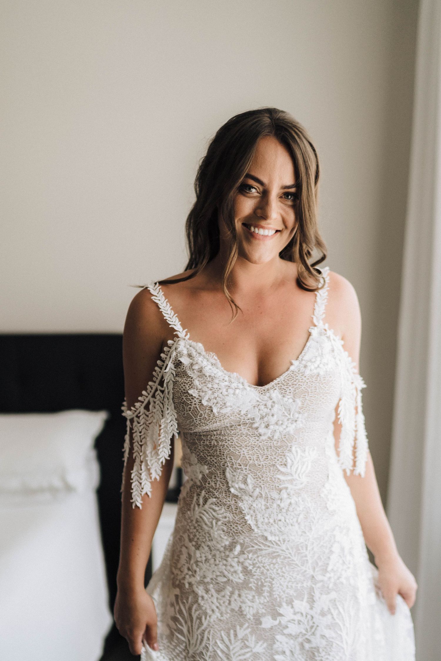 A bride wearing a delicate lace wedding dress with cold shoulder details smiles warmly in a bright room.