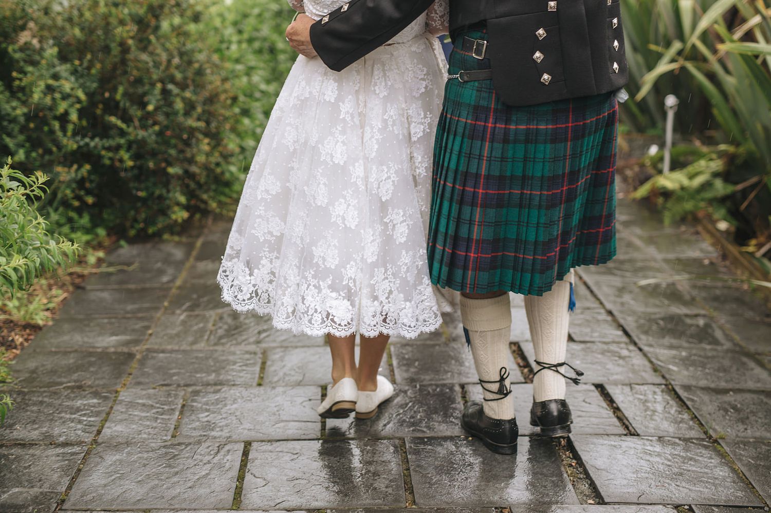 White lace wedding dress and green plaid kilt with traditional Scottish socks visible from knees down on wet stone path.