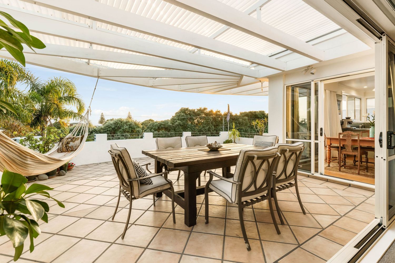 Covered outdoor patio area with dining table and chairs overlooking landscaped garden under white pergola structure.