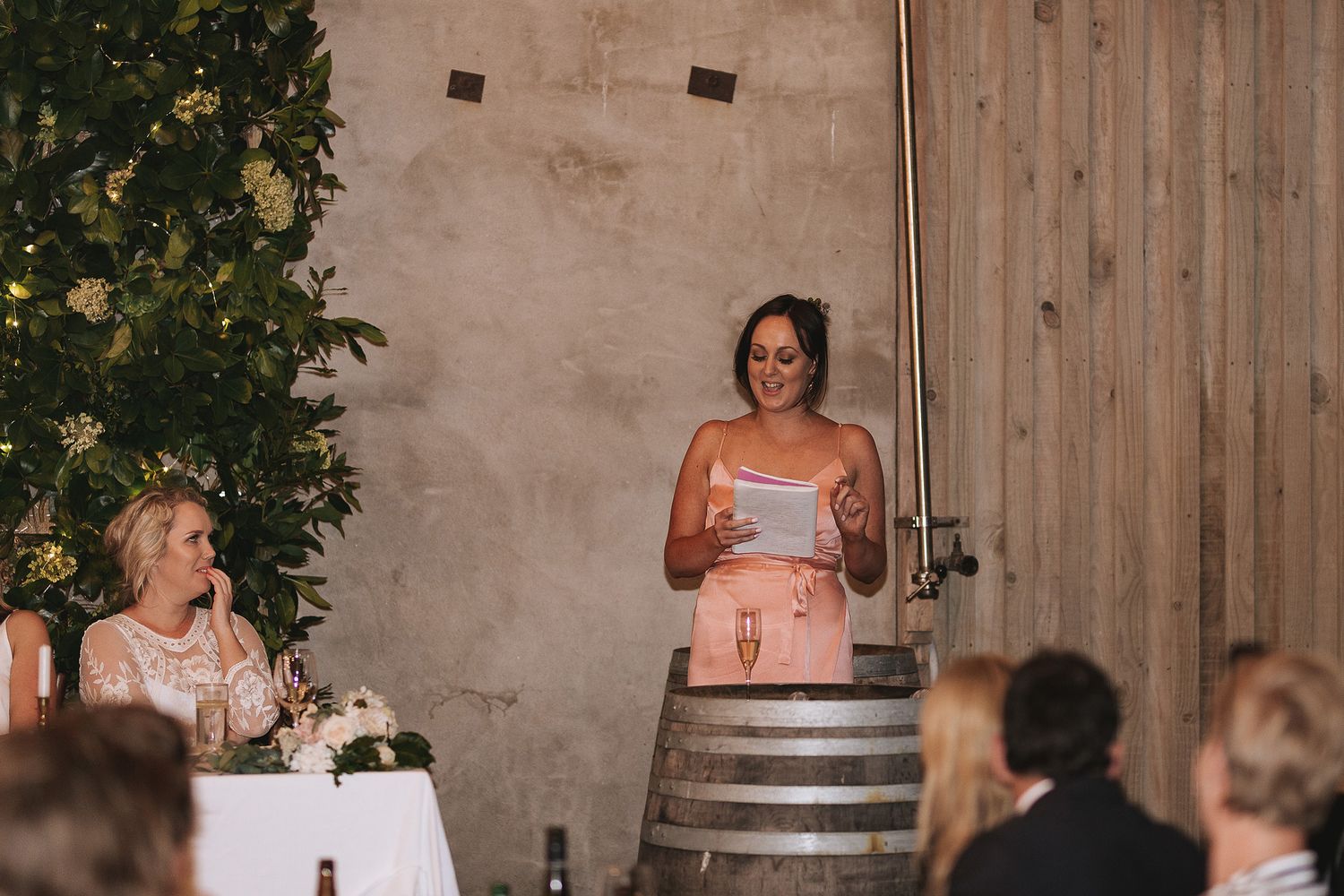 Guests stand and applaud during a winery wedding reception with wine barrels and rustic wall decor in background.
