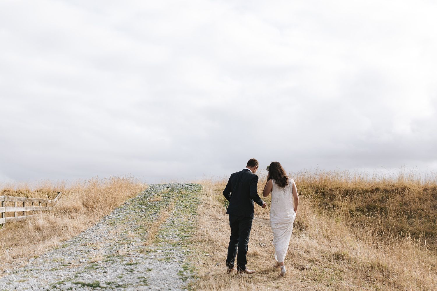 Two figures walk together on a winding dirt path through dried grass fields under an overcast sky.