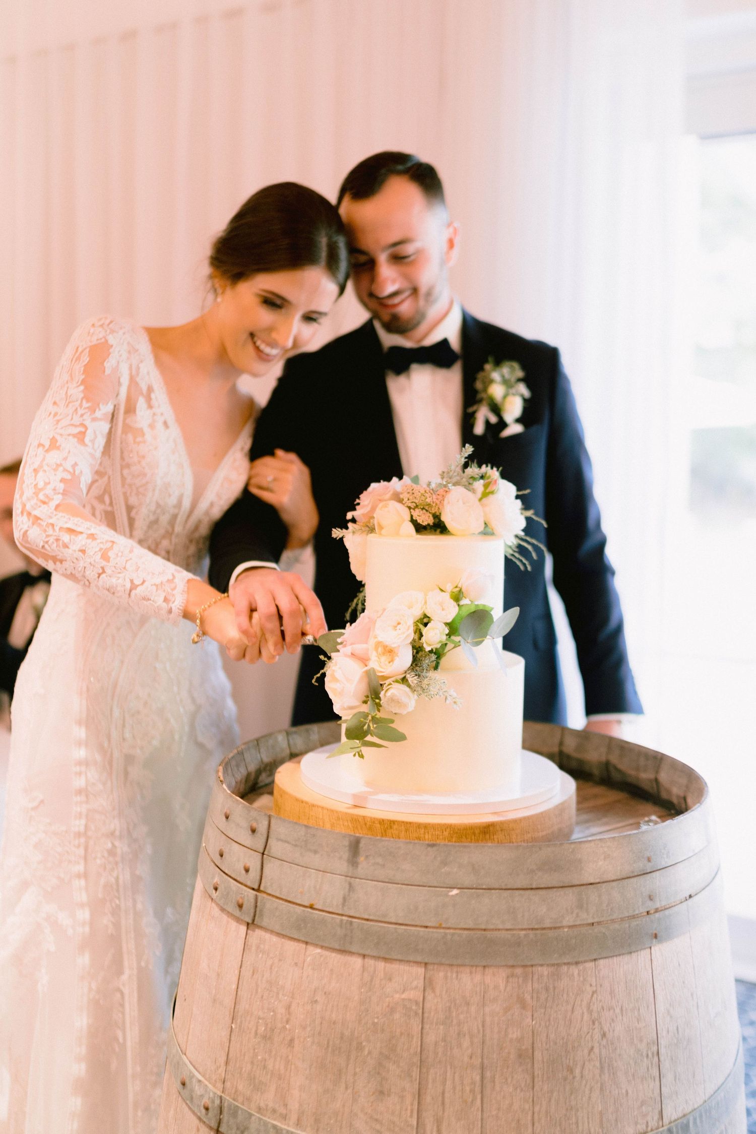 Newlyweds cutting their elegant white wedding cake displayed on a rustic wine barrel.