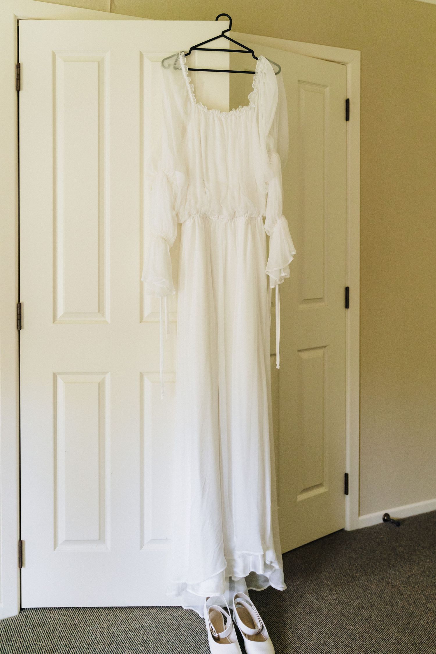 An elegant white wedding dress and shoes displayed on a hanger against a white door.