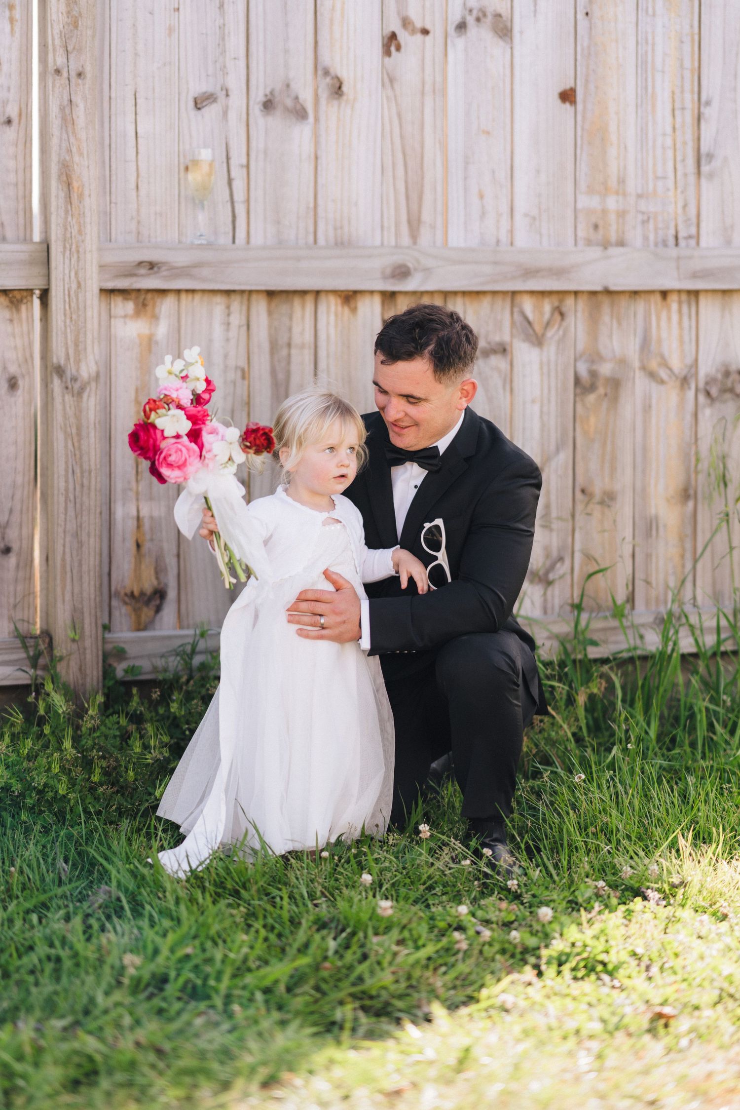 A sweet moment in front of a wooden fence, with a bouquet of pink flowers adding a pop of color against the rustic backdrop.