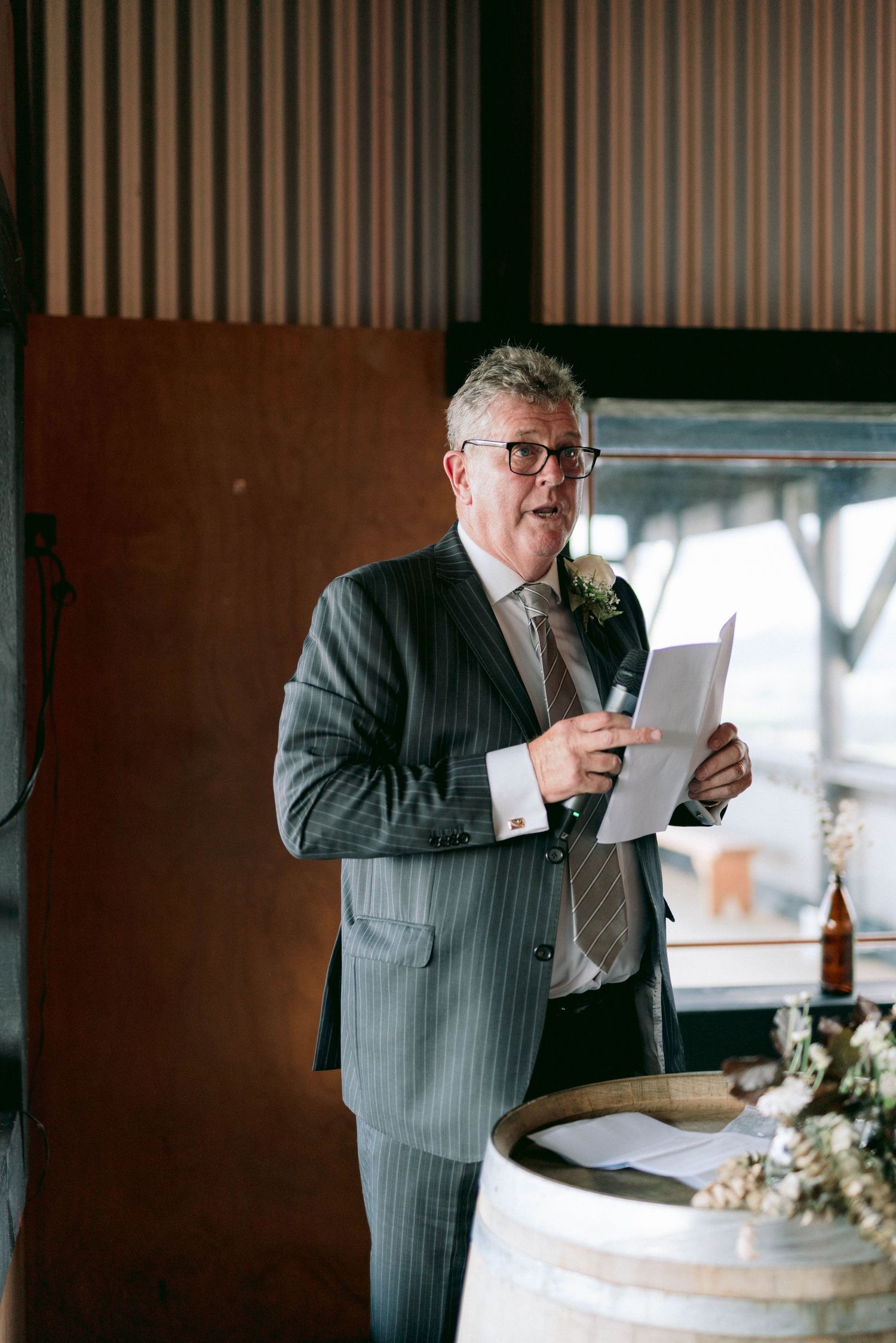 A person in a pinstriped suit reads from papers while standing at a wooden wine barrel podium.