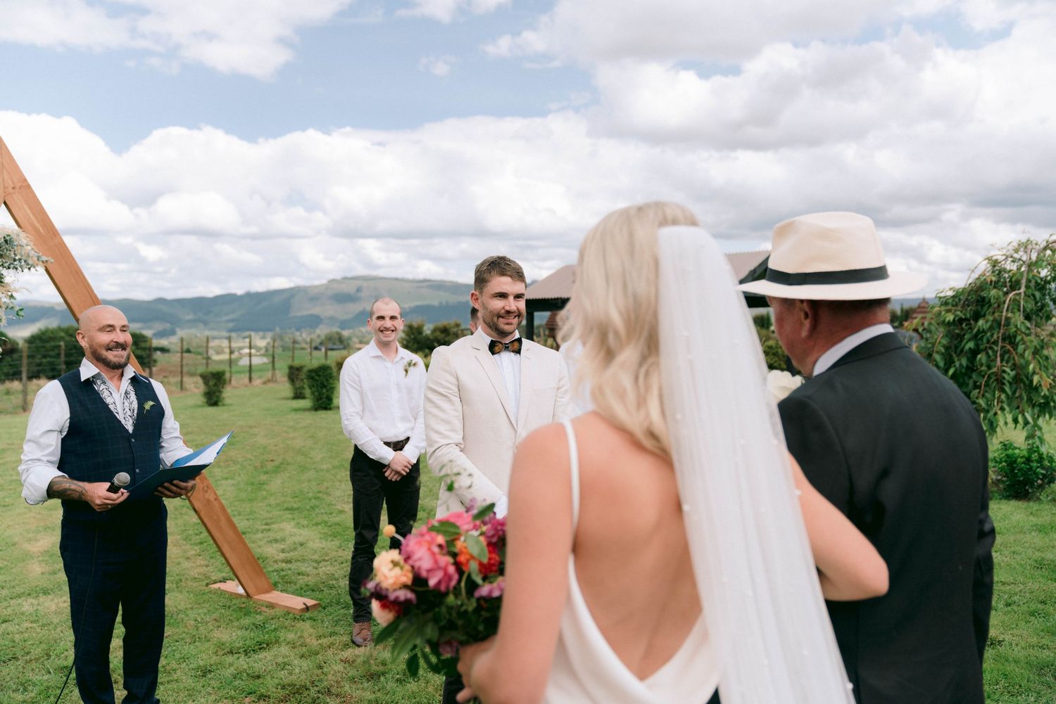 An outdoor wedding ceremony takes place with guests gathered on a scenic hillside overlooking mountains.
