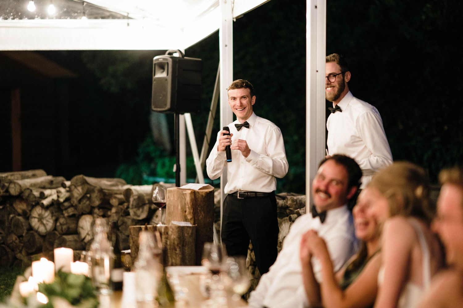 Evening wedding reception with speakers giving toasts under string lights.