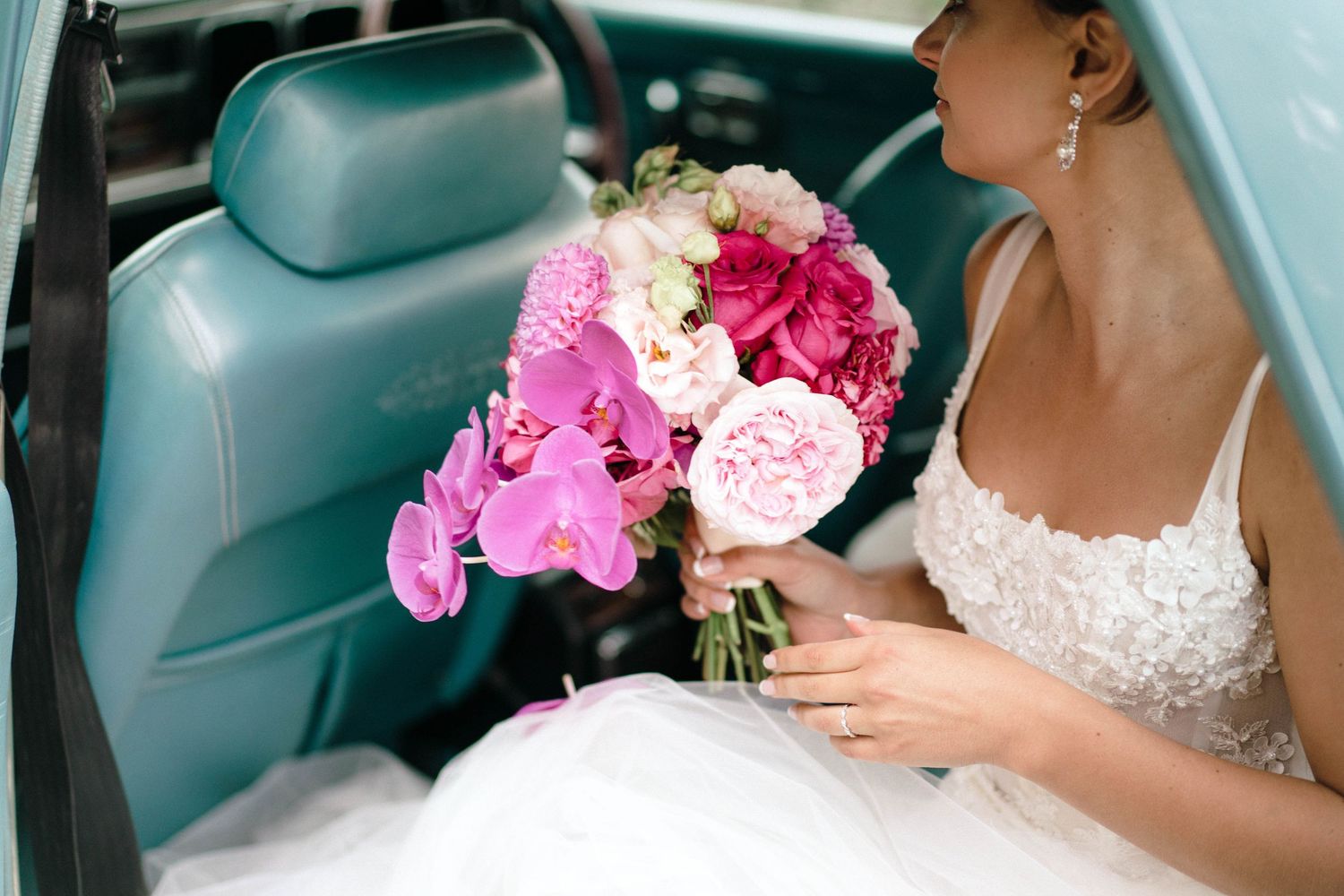 Pink and fuchsia wedding bouquet held by someone wearing a white lace dress in a car with teal seats.