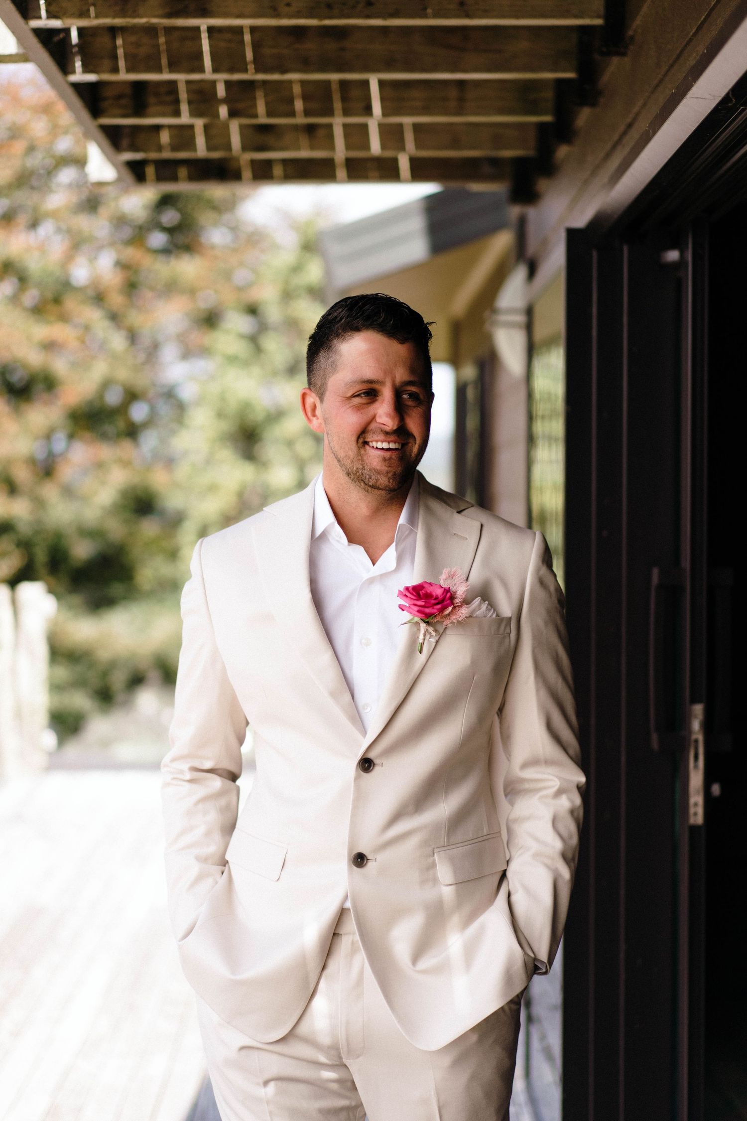 Groom in cream-colored suit with pink boutonniere poses on porch with wooden beams.