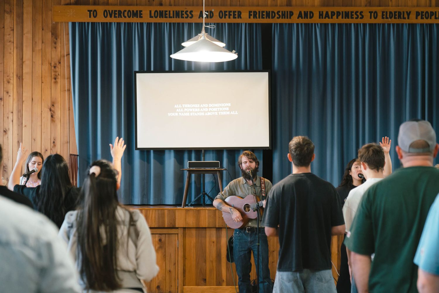 Musicians performing for an audience in a wood-paneled room with projection screen.