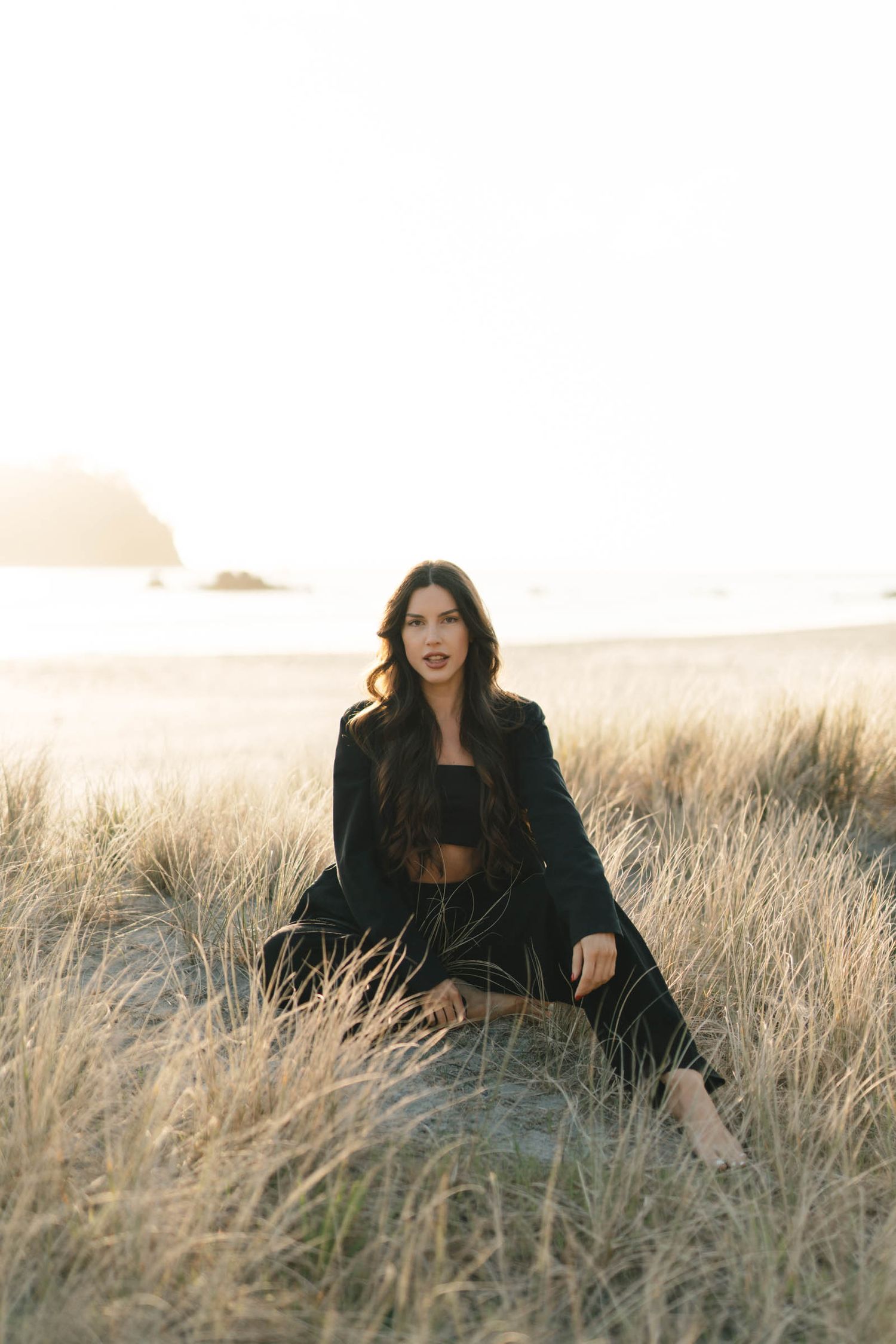 A person in dark clothing sits playfully in tall grass with coastal scenery behind them.