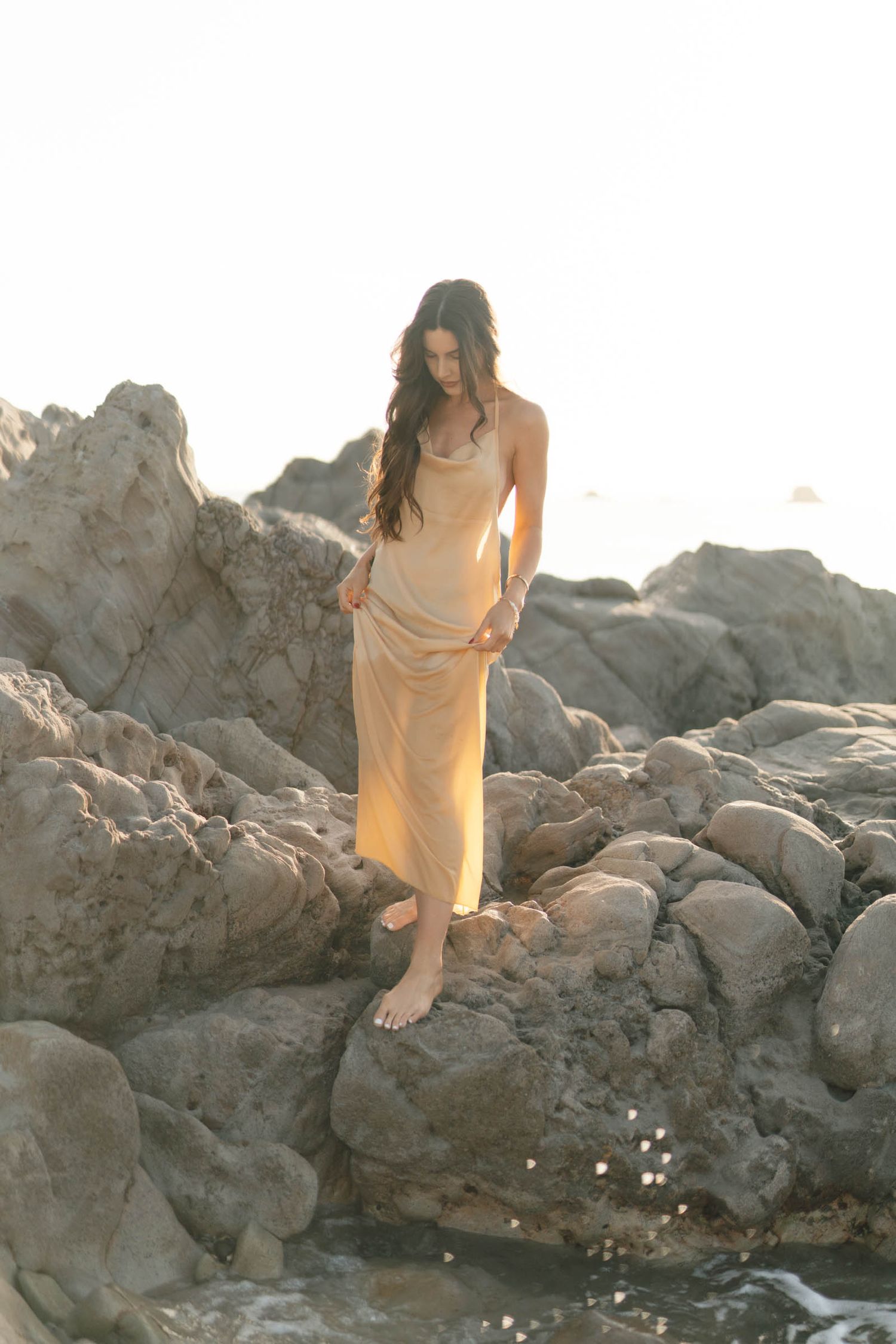 A feminine figure in a light dress stands gracefully on coastal rocks during sunset.