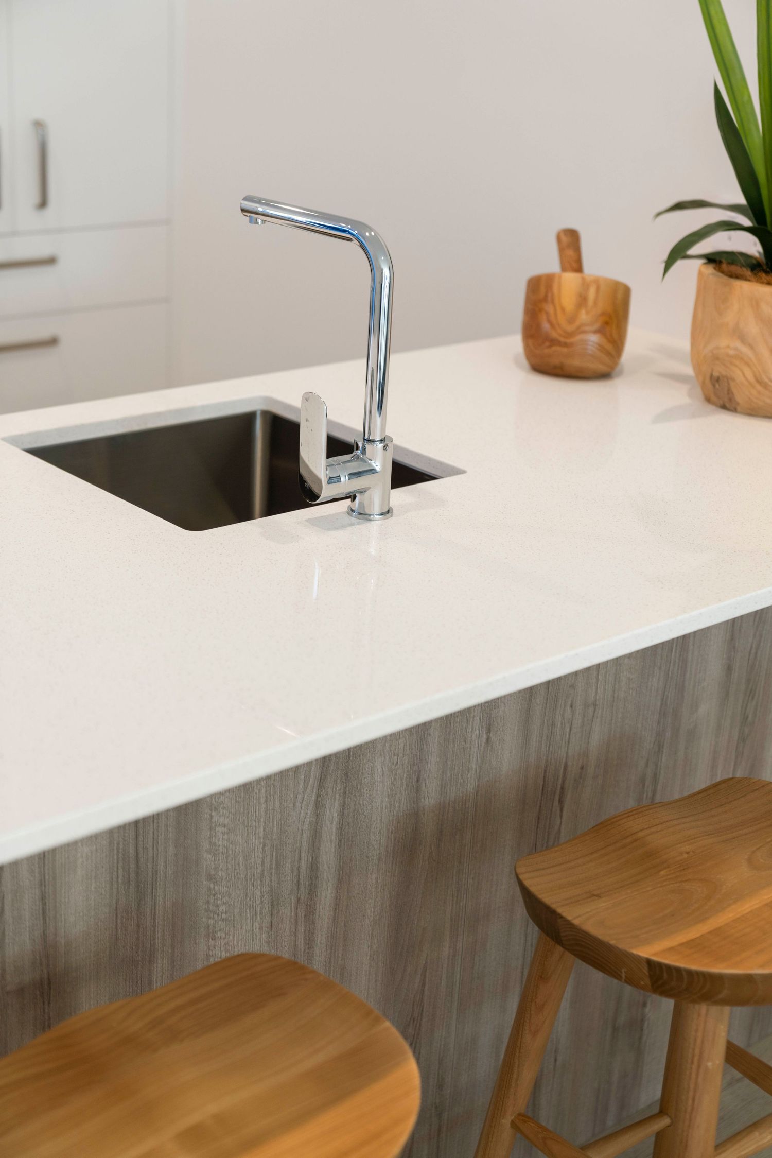 White kitchen counter with wooden bar stools and sleek chrome faucet against grey backsplash.