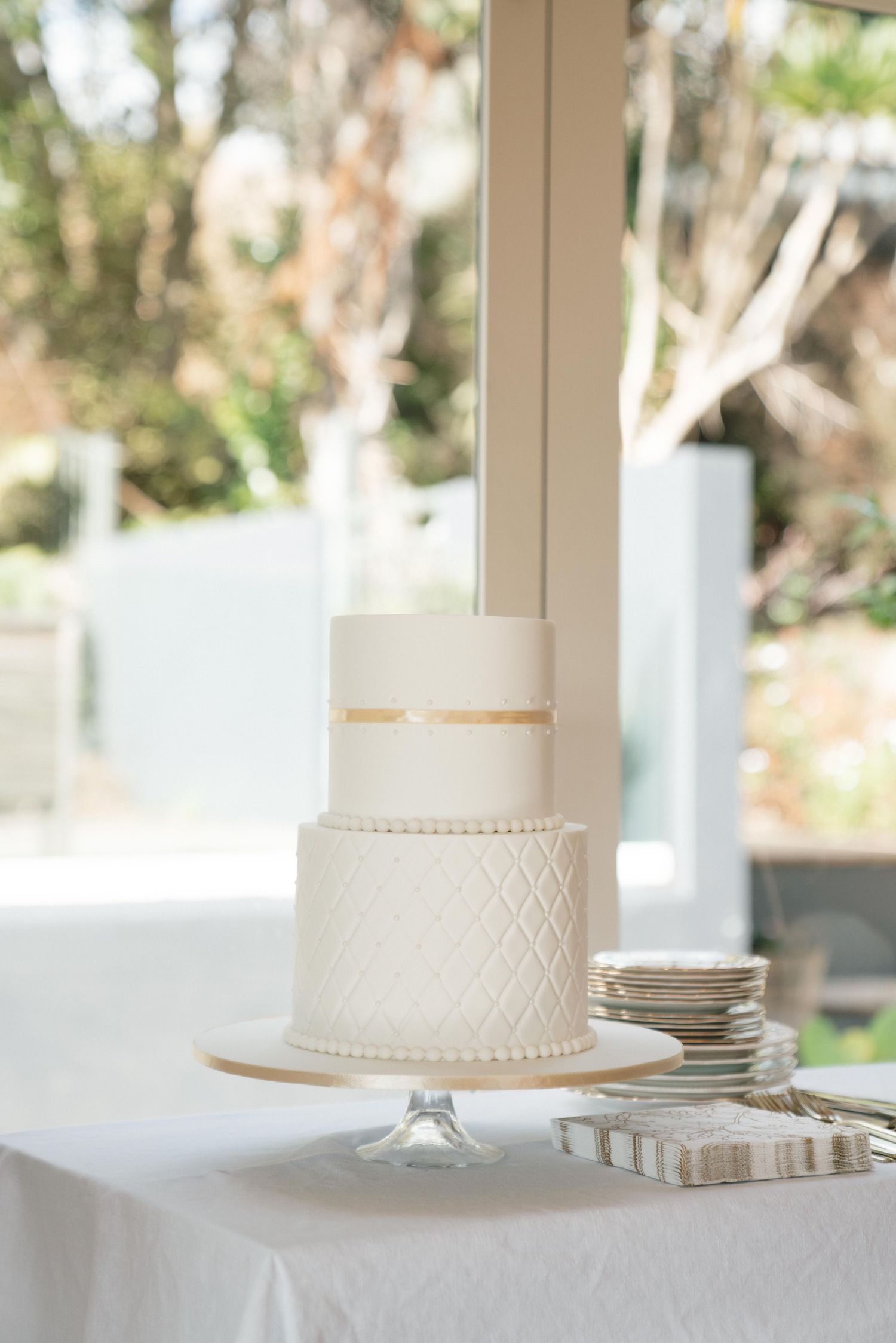 White wedding cake and plates displayed on an elegant white tablecloth near sunny windows.