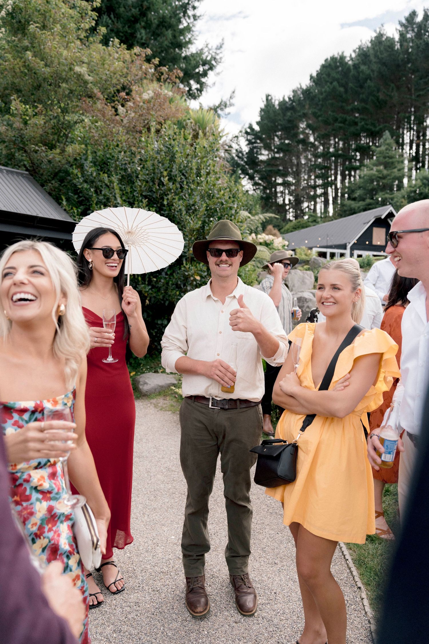 Wedding guests mingle outdoors in summer attire with parasol and cocktails near pine trees.