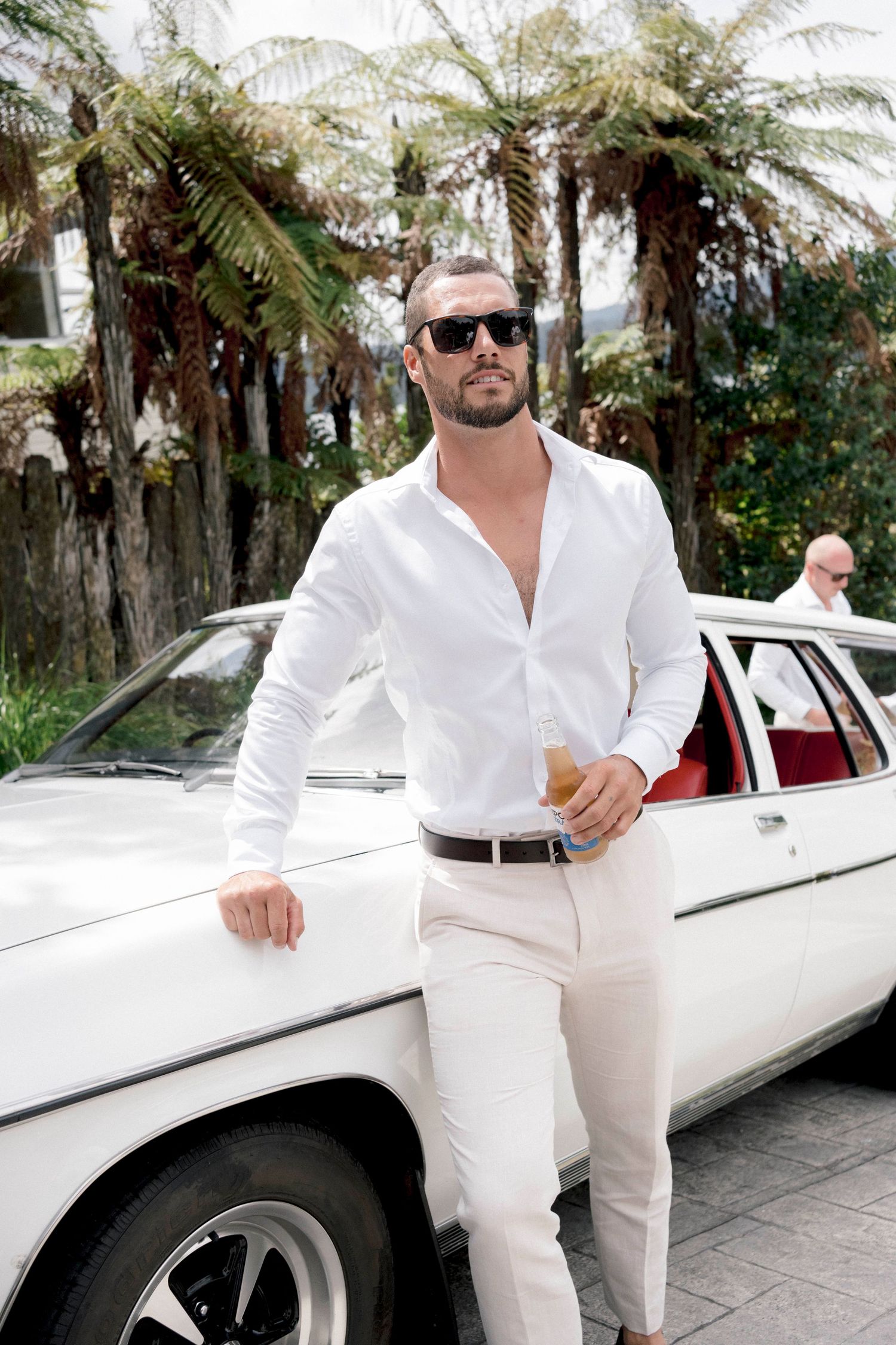 A person in all-white clothing poses next to a classic white limousine surrounded by palm trees.