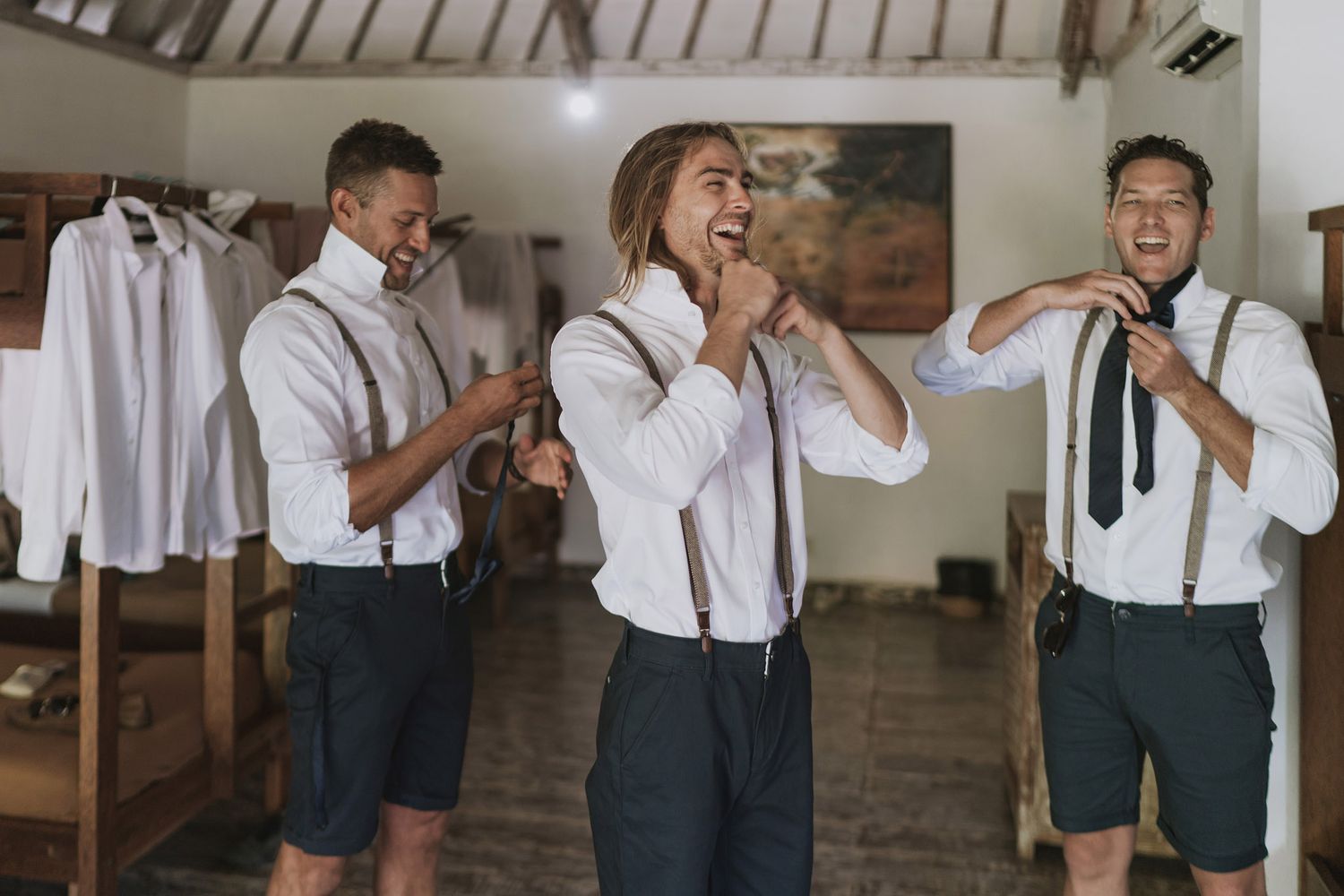 Wedding preparation scene showing people getting dressed in a rustic room with white shirts and suspenders.