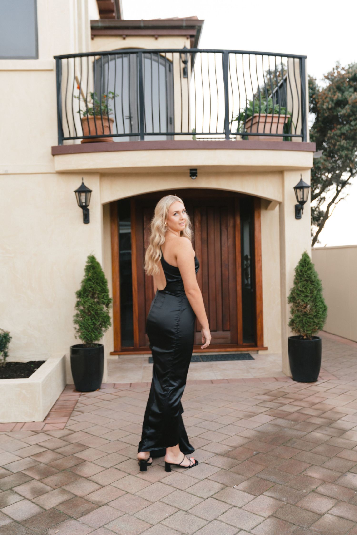 Woman in elegant black dress poses in front of Mediterranean-style home entrance with balcony.