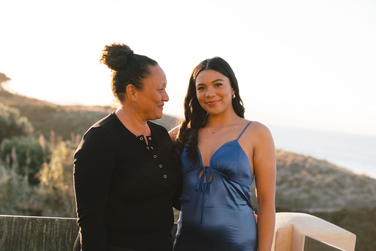 Two people share a warm moment together during golden hour on a scenic hillside overlooking the ocean.