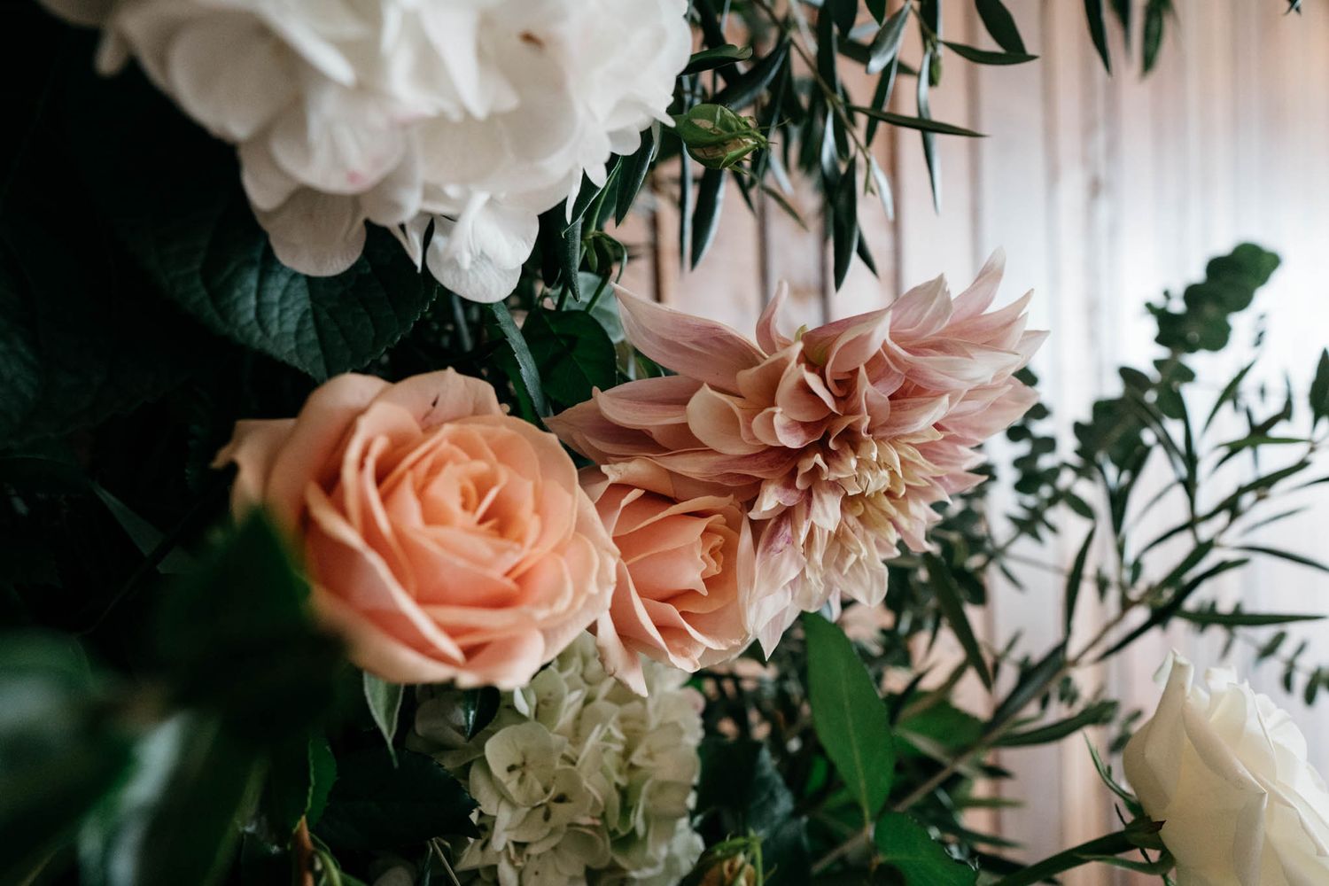 Close-up of peach roses and blush dahlias with white hydrangeas and green foliage in elegant floral arrangement.