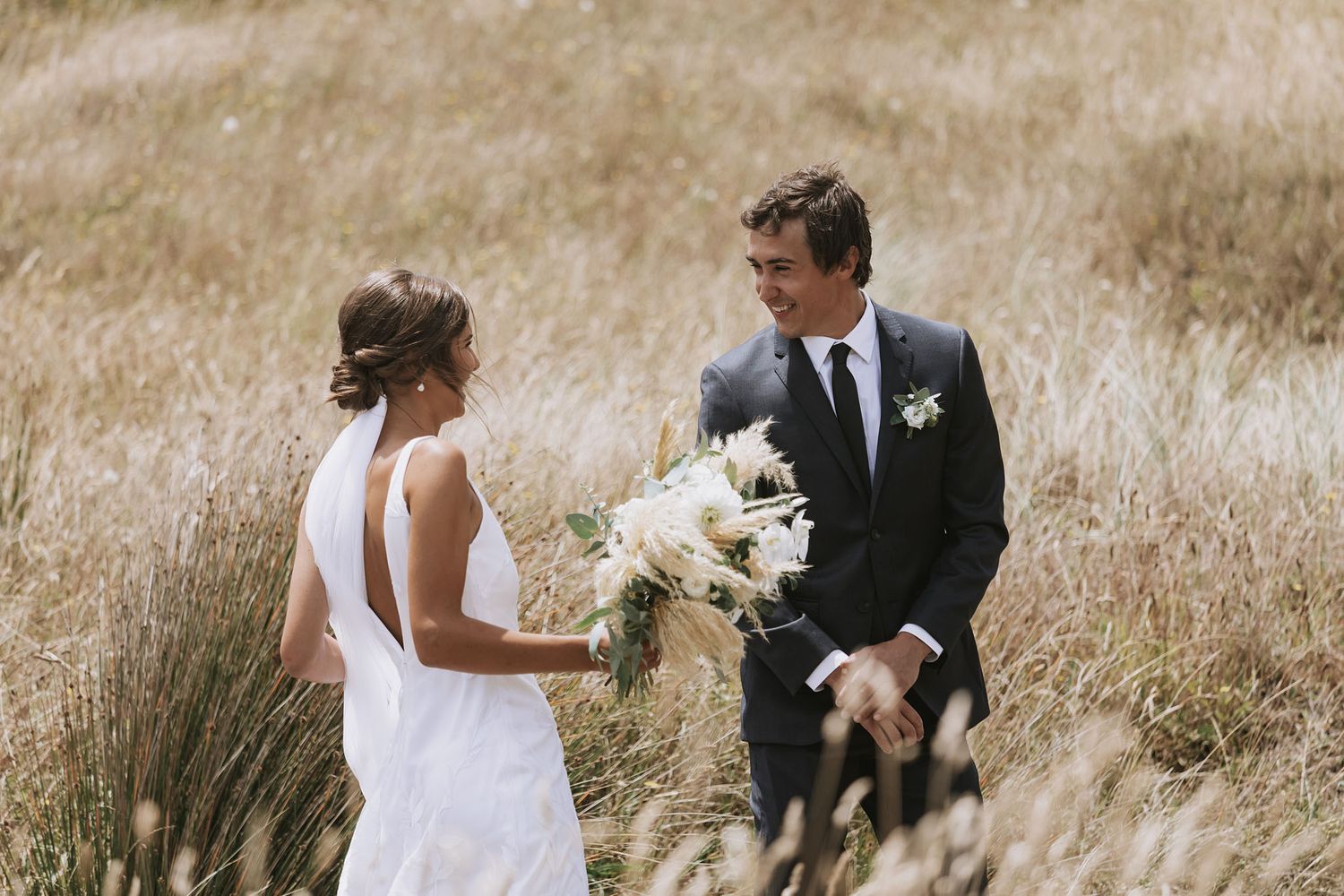 Newlywed couple walks hand in hand through a golden field with bridal bouquet and formal wedding attire.
