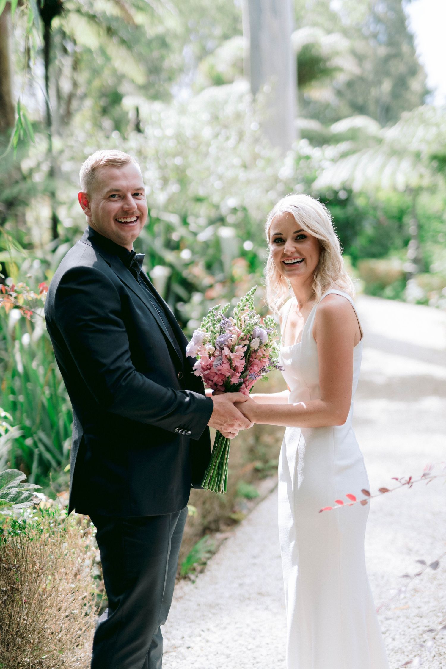 A bride in a white dress and groom in black suit share a joyful moment while holding a pink bouquet in a garden setting.