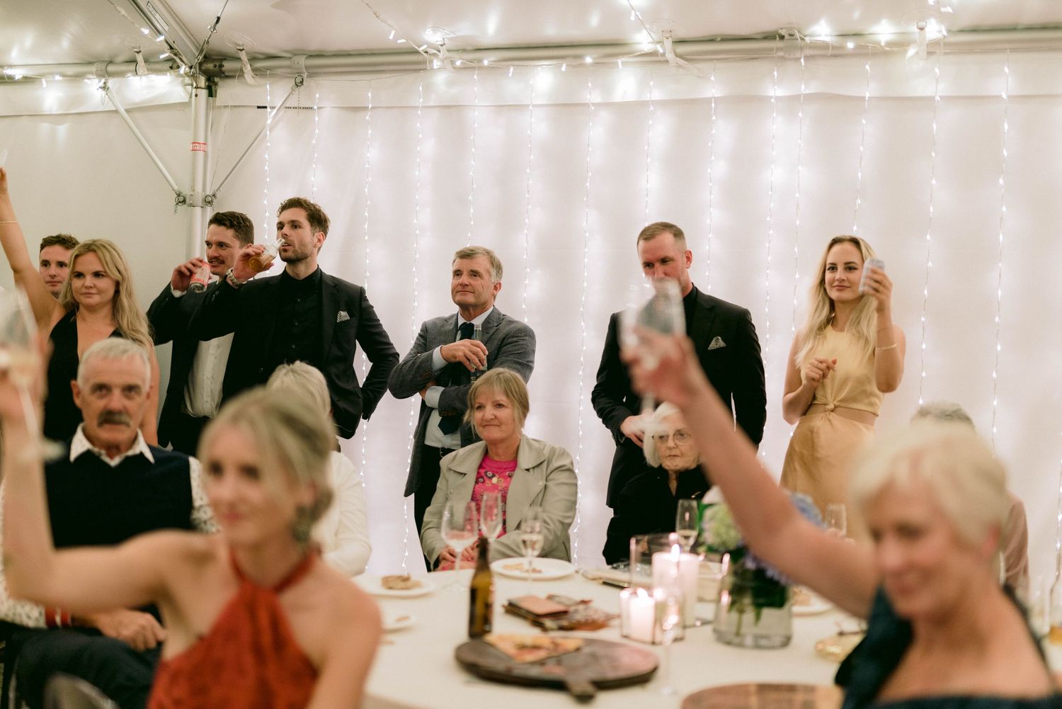 Wedding guests enjoying a celebratory reception with string lights and decorations in an indoor venue.