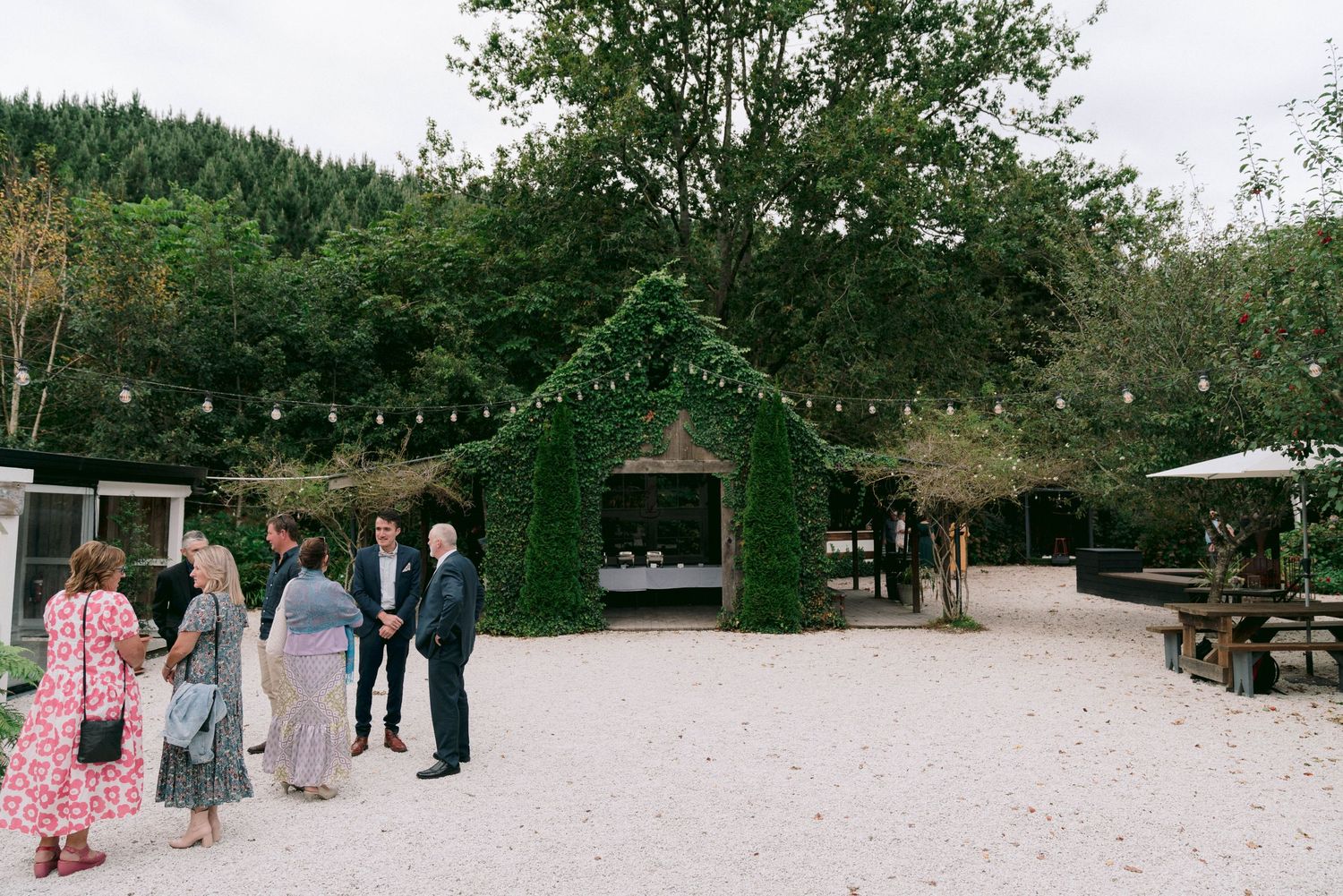 An ivy-covered archway with string lights in a garden setting where guests gather on the gravel path.