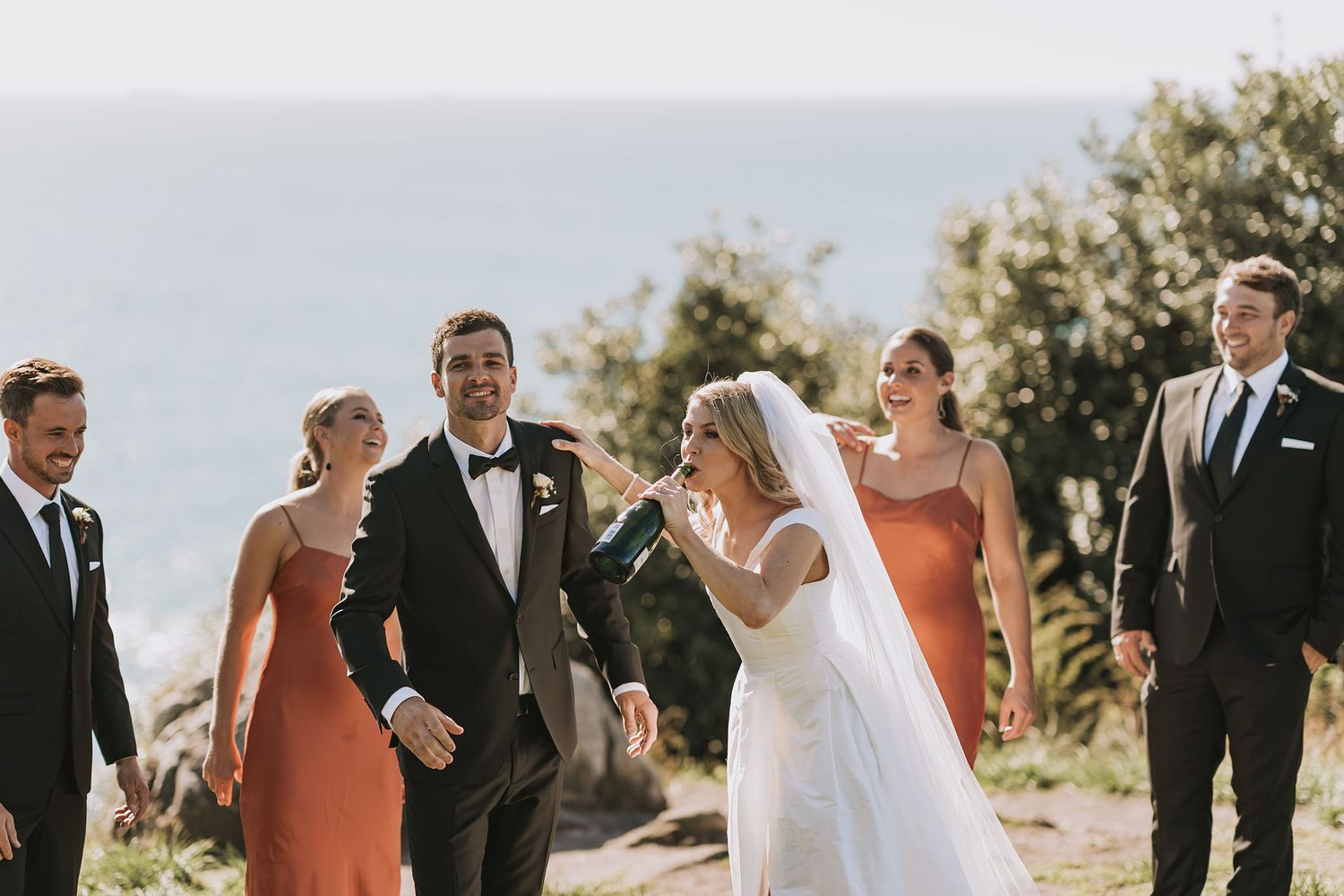 Wedding party laughs together during outdoor ceremony on sunny mountainside with bridesmaids in coral dresses.