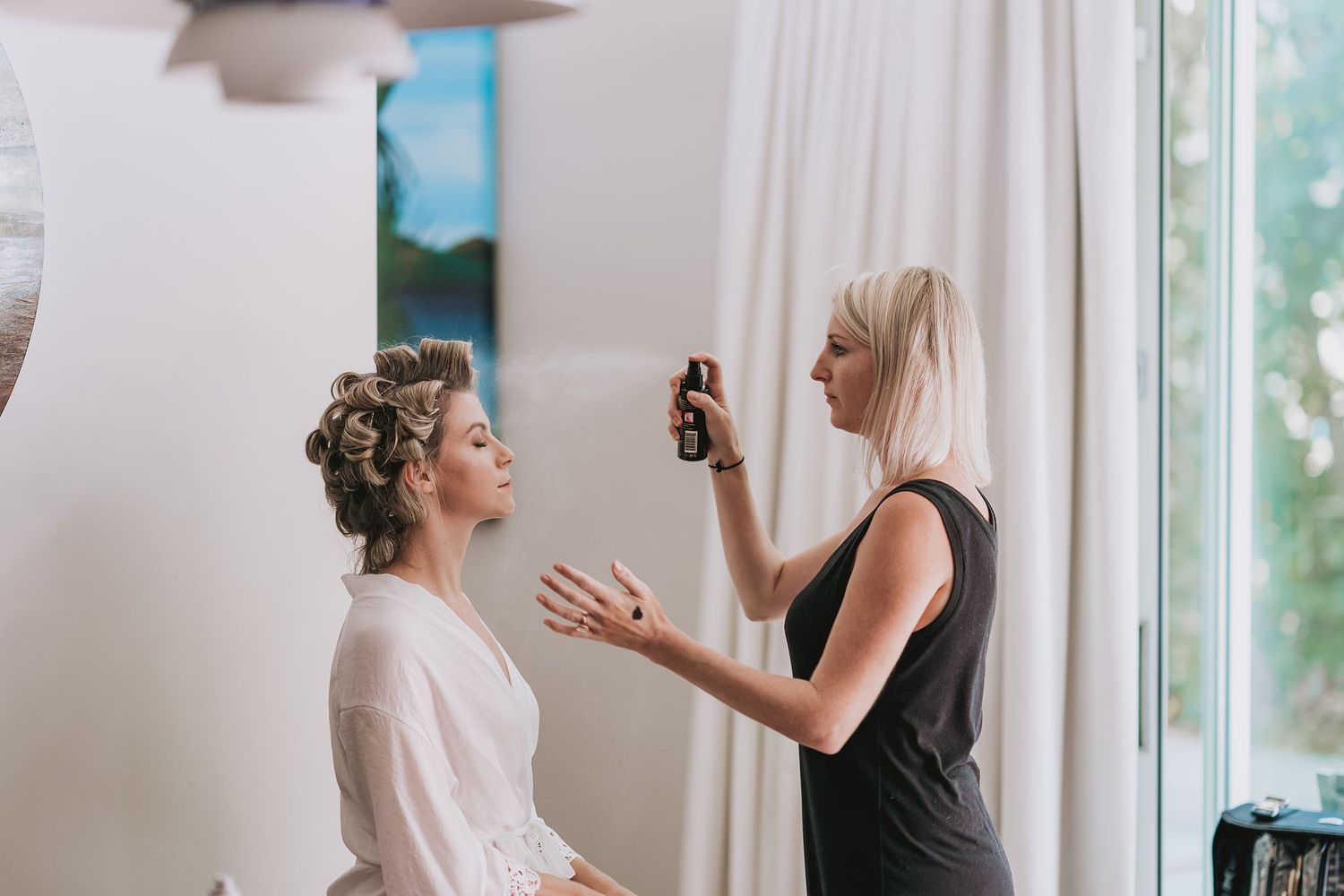 Professional hairstylist spraying hairspray while finishing bridal hair preparation near window.
