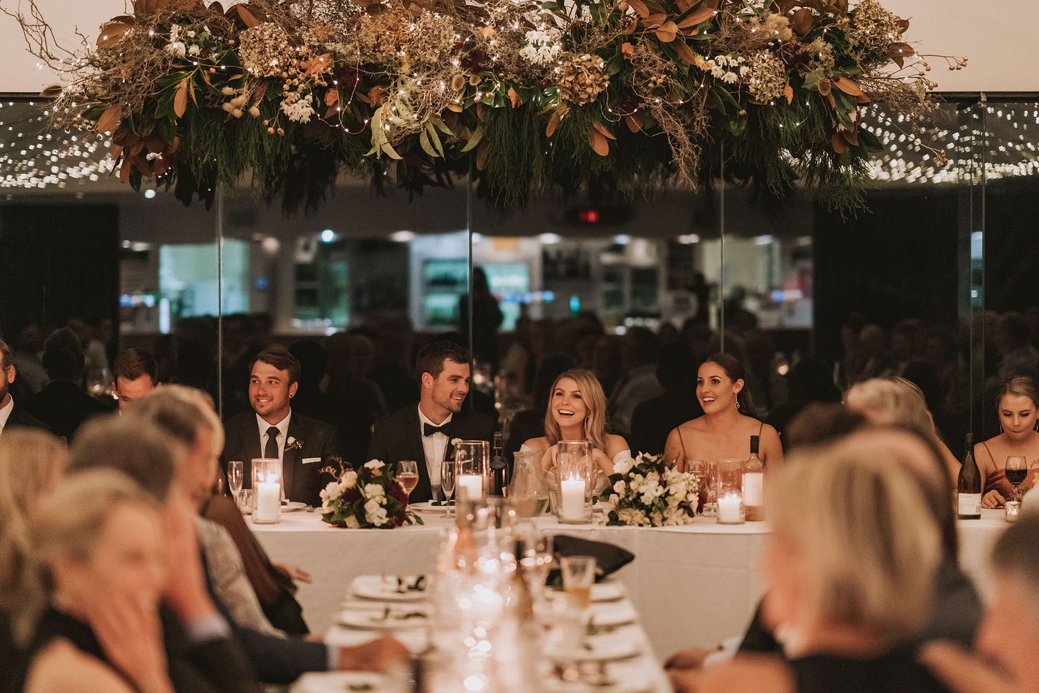 Wedding reception dinner setup with elegant floral arrangements and decorative lighting overhead.