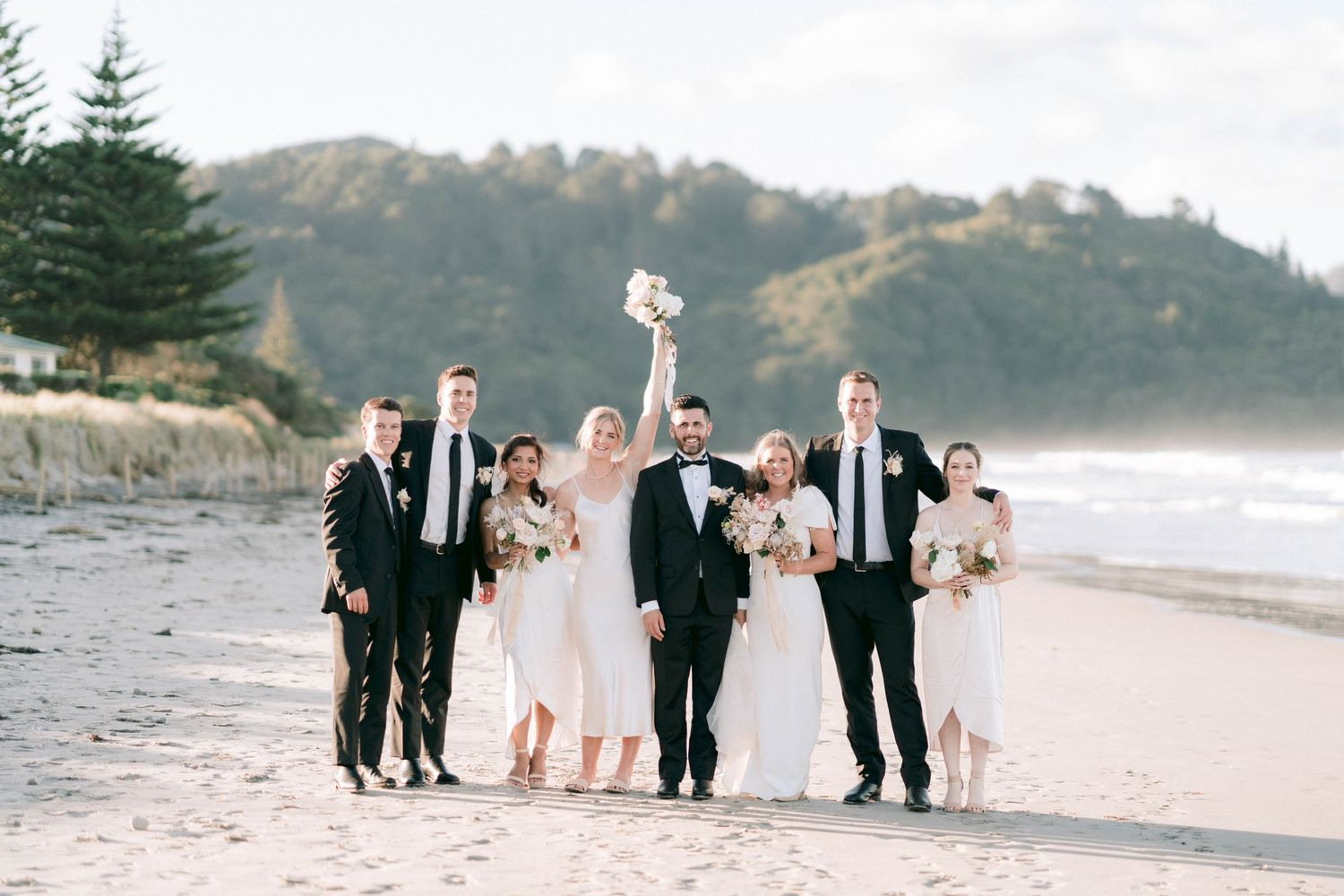 Wedding party poses together on scenic beach with mountains and evergreen trees in background.