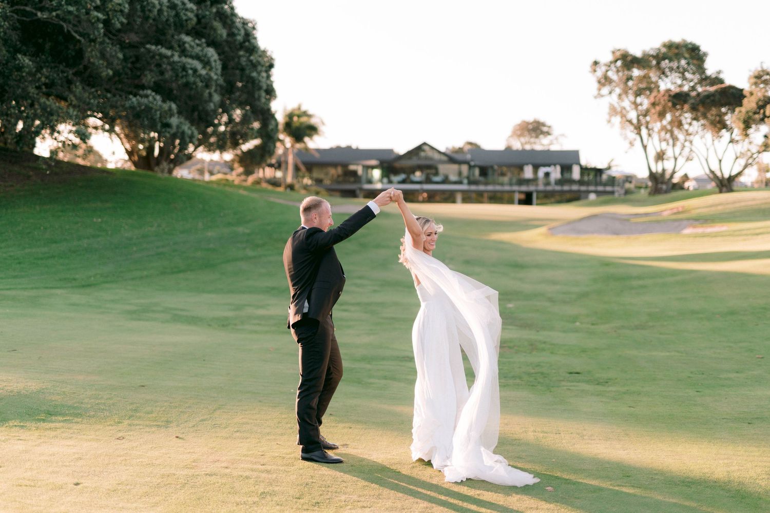 Two people strike joyful poses on a lush golf course at sunset with buildings in the background.
