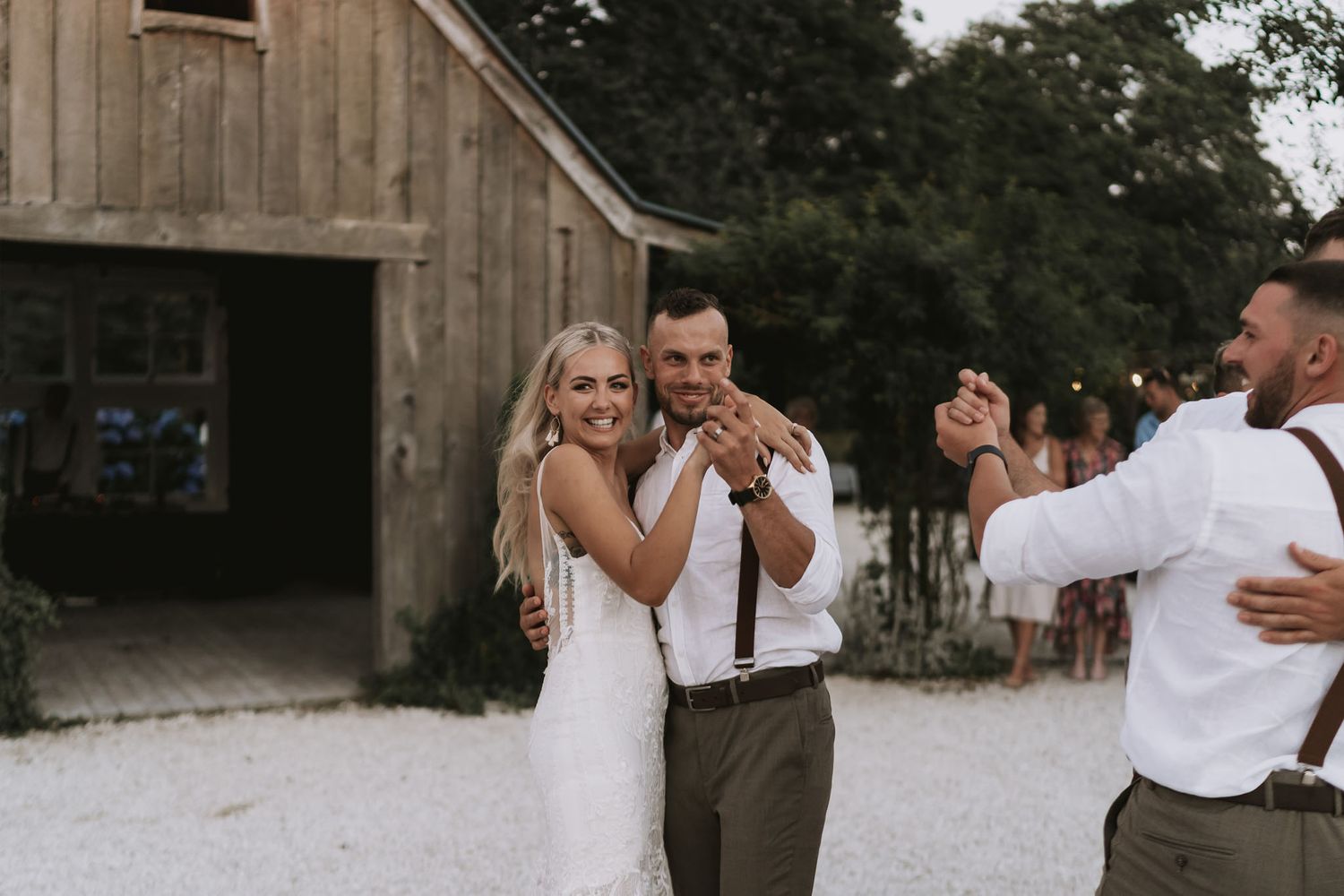 Wedding party dancing outdoors at rustic barn venue with string lights and casual white attire at dusk.