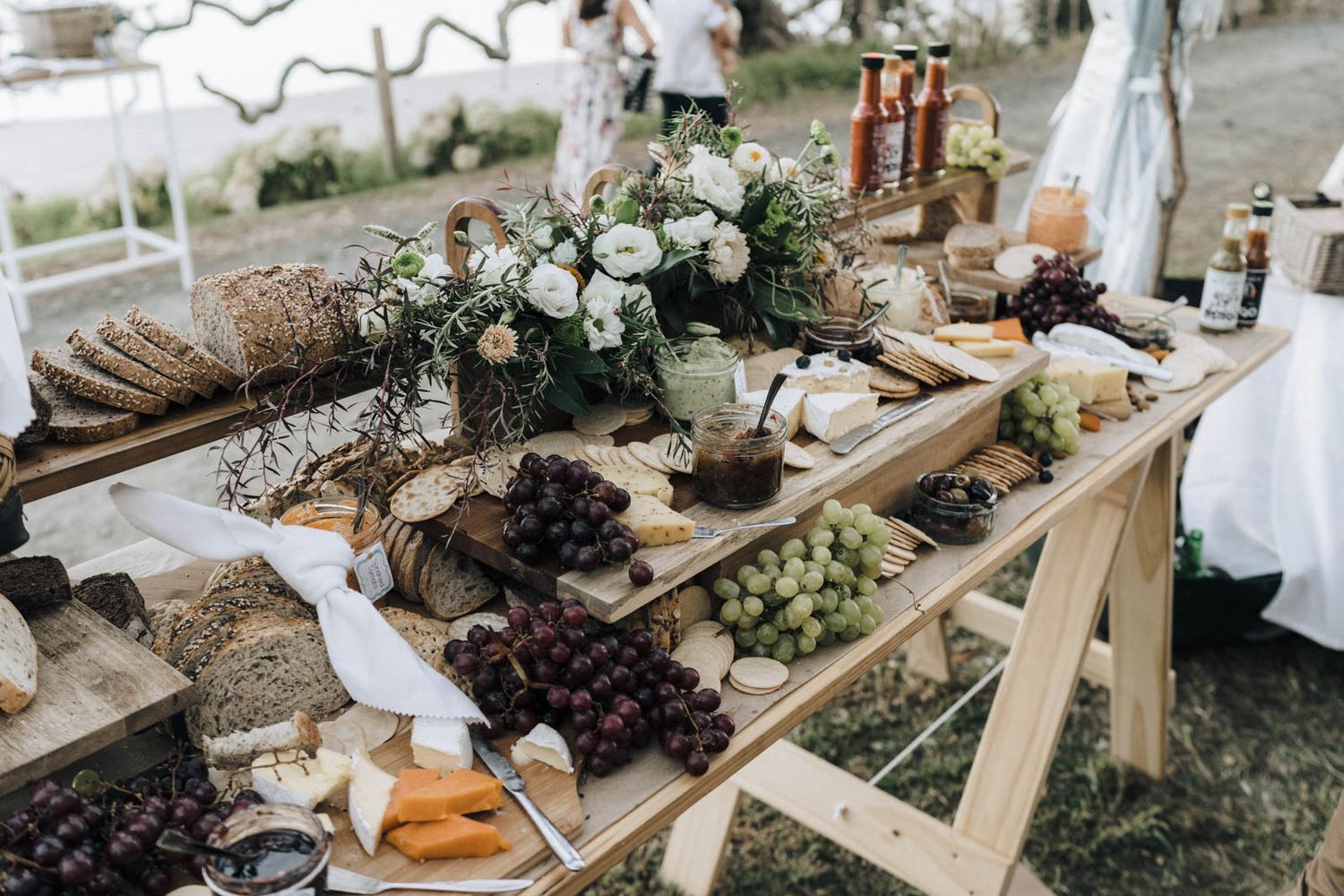Rustic outdoor wedding grazing table decorated with greenery, cheese, bread, grapes and floral arrangements.