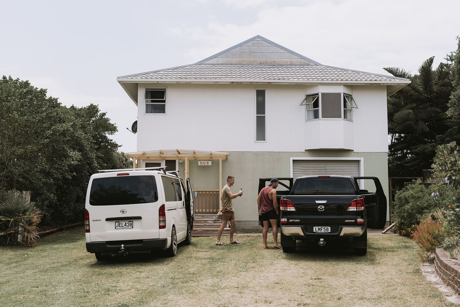 A white two-story beach house with carport and parked vehicles on a grassy property.