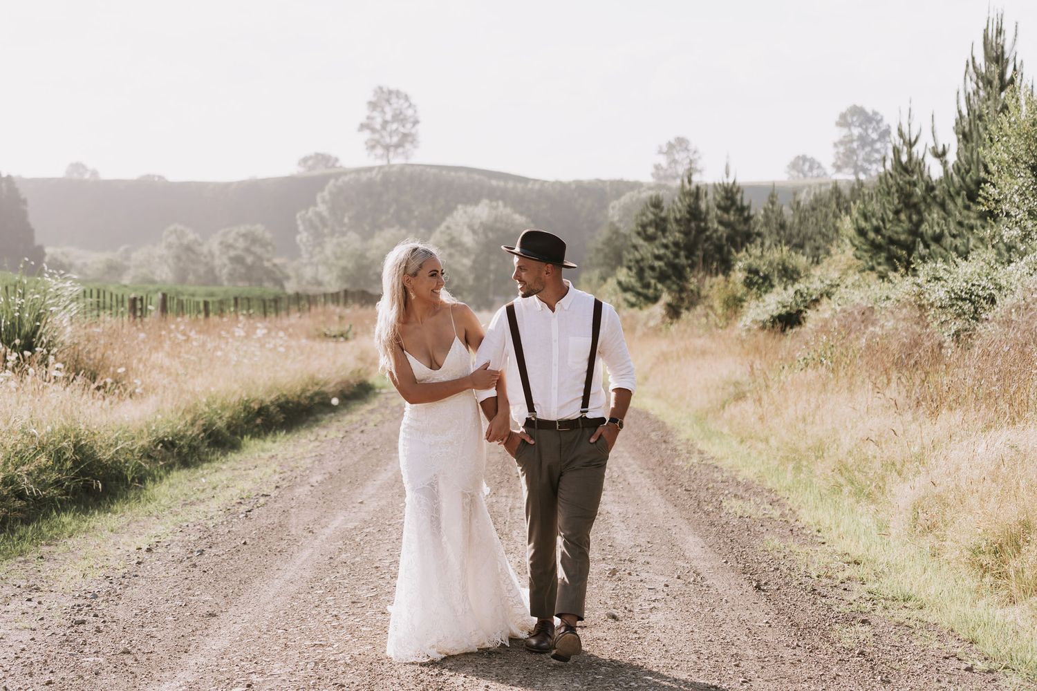 A newlywed couple walks hand in hand down a dusty country road through golden fields on their wedding day.