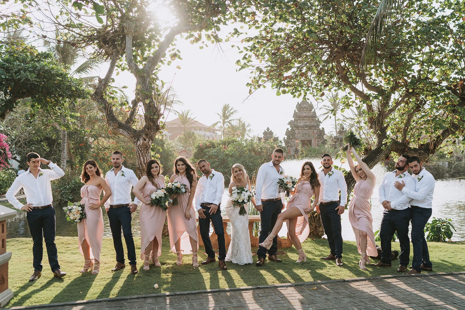 A wedding party jumps in celebration on a sunny lawn with trees in the background.