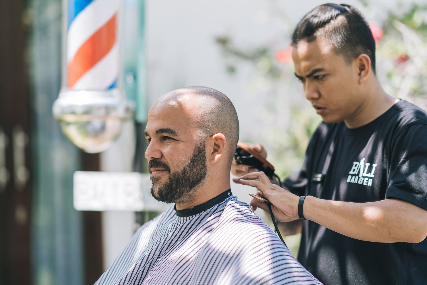 Professional barber trims client's hair at traditional barbershop with classic striped pole visible outside.