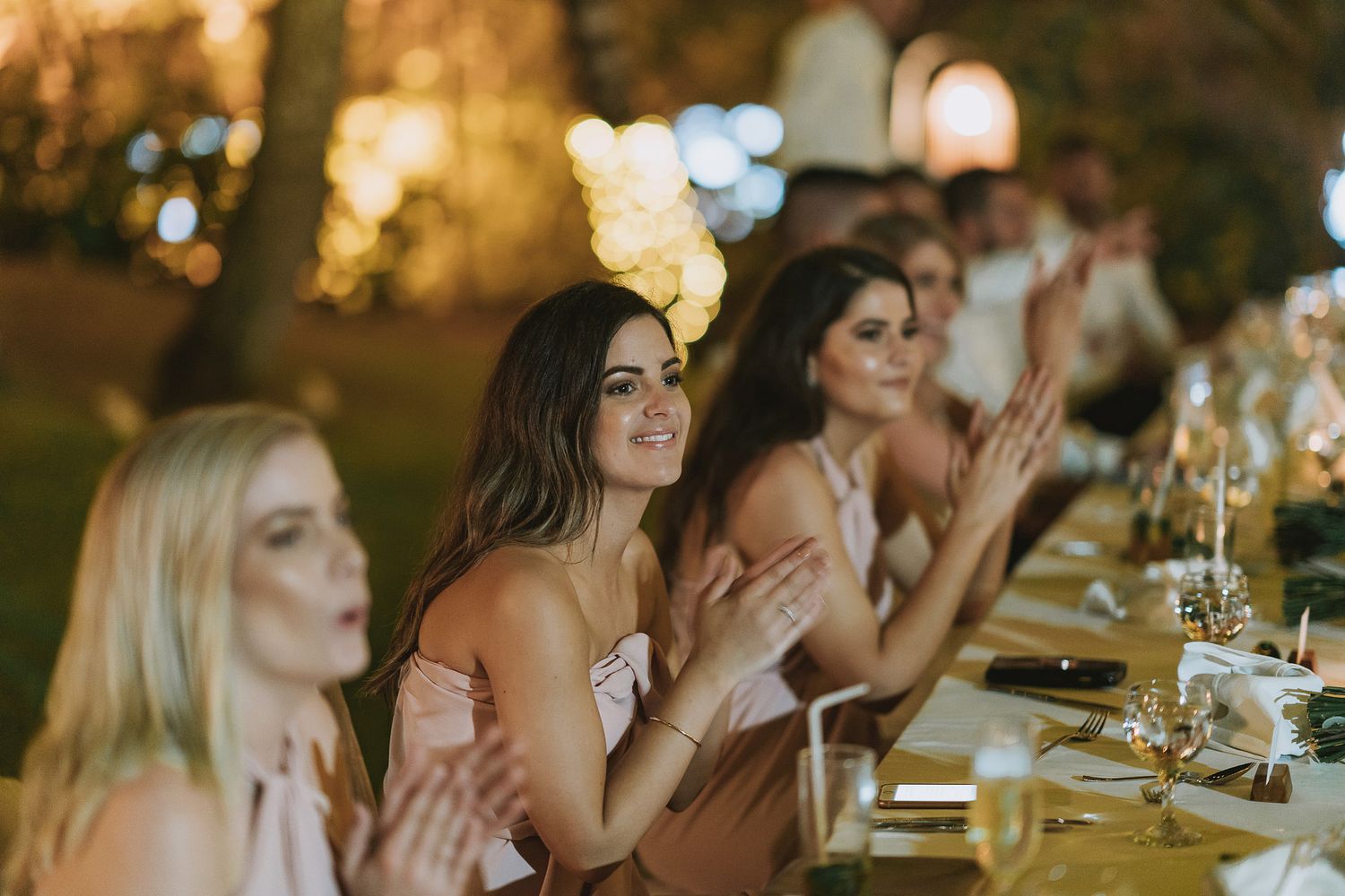 Wedding guests applauding during an outdoor reception dinner under twinkling lights.
